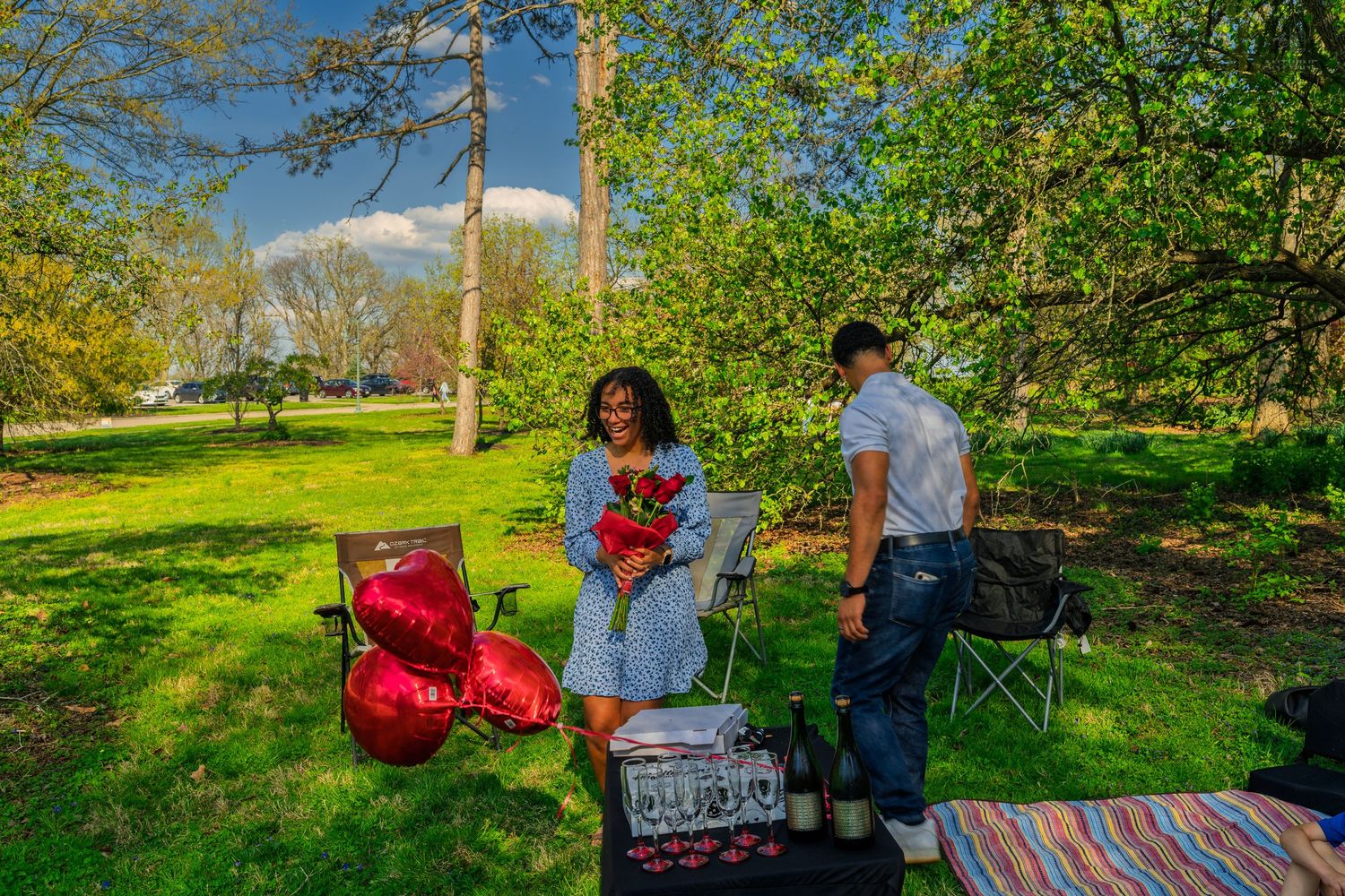 Couple sharing a romantic outdoor picnic proposal setup with red heart balloons, roses, and champagne in a park.