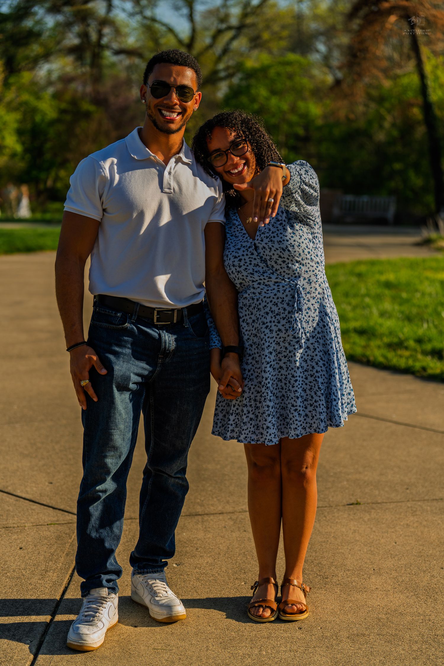 Smiling couple posing together in a sunny park, man in white polo and woman in blue floral dress.
