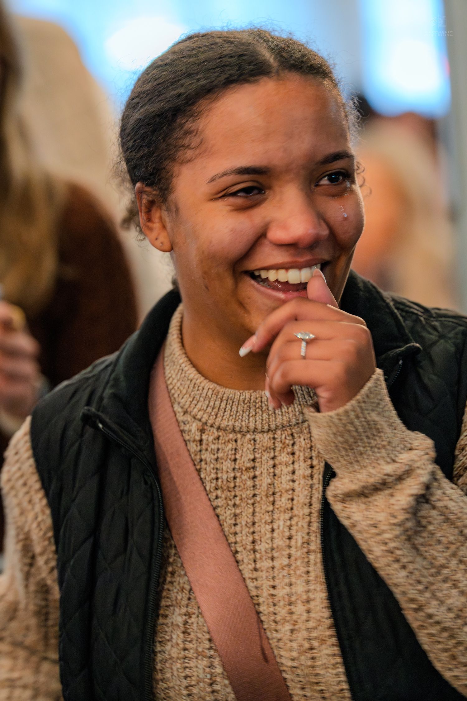 A smiling young woman in a beige sweater and black vest enjoys a cheerful moment at a social event.