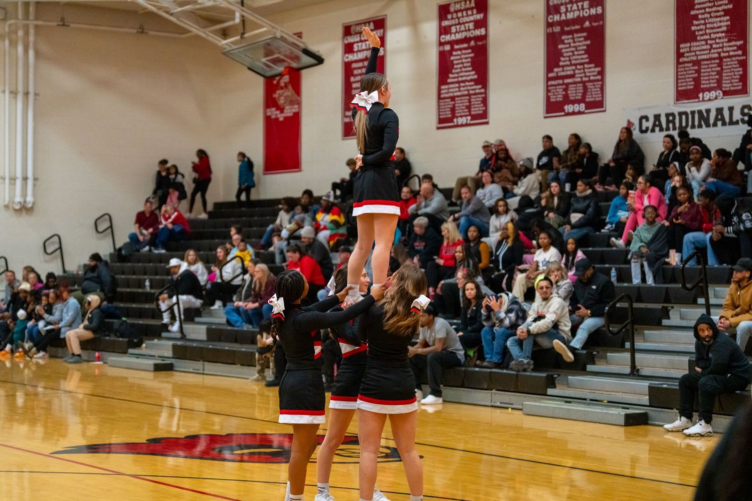 High school cheerleaders perform a stunt with one cheerleader lifted high during a packed basketball game.