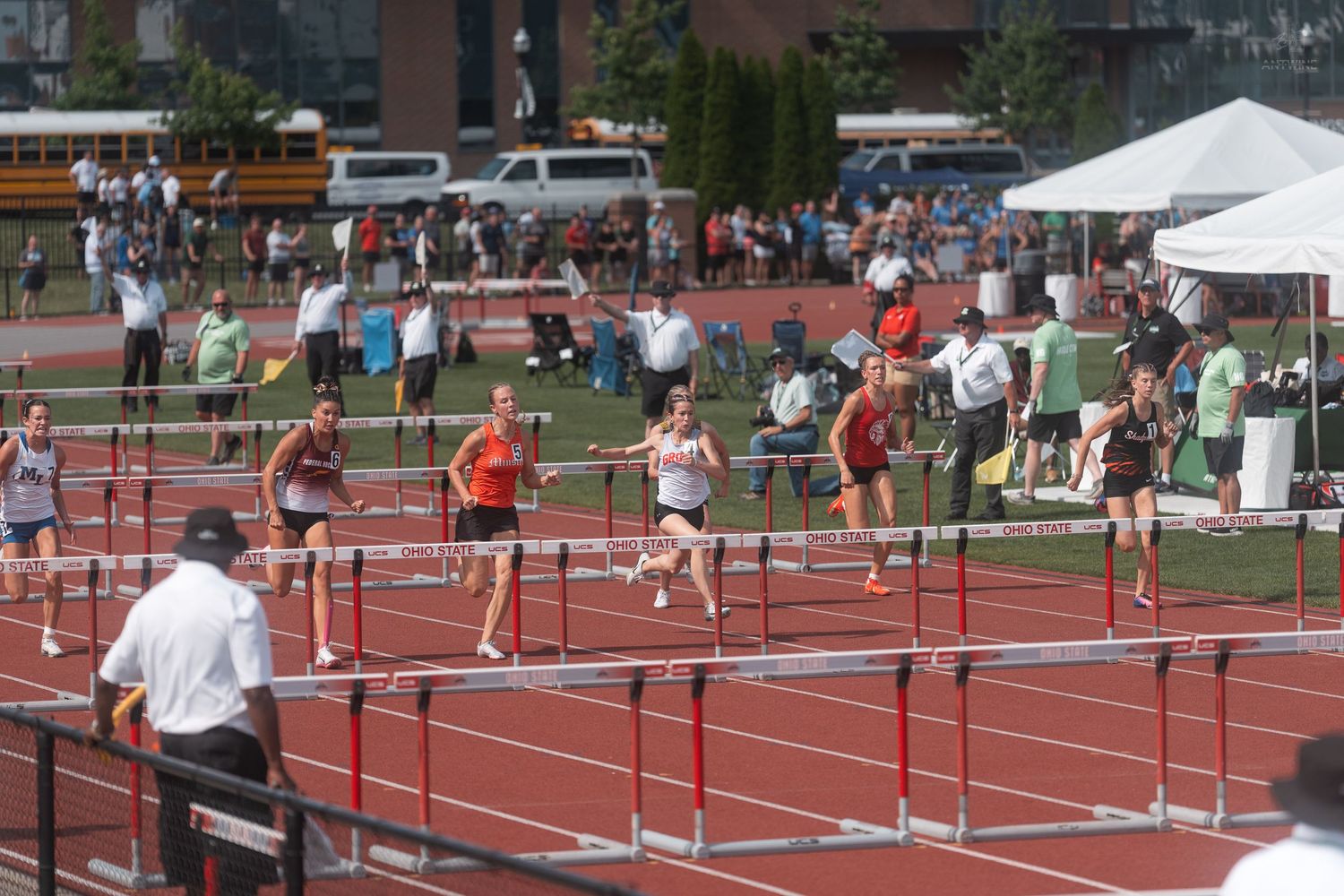 Track athletes race over hurdles on red track with official observing during outdoor athletics competition.