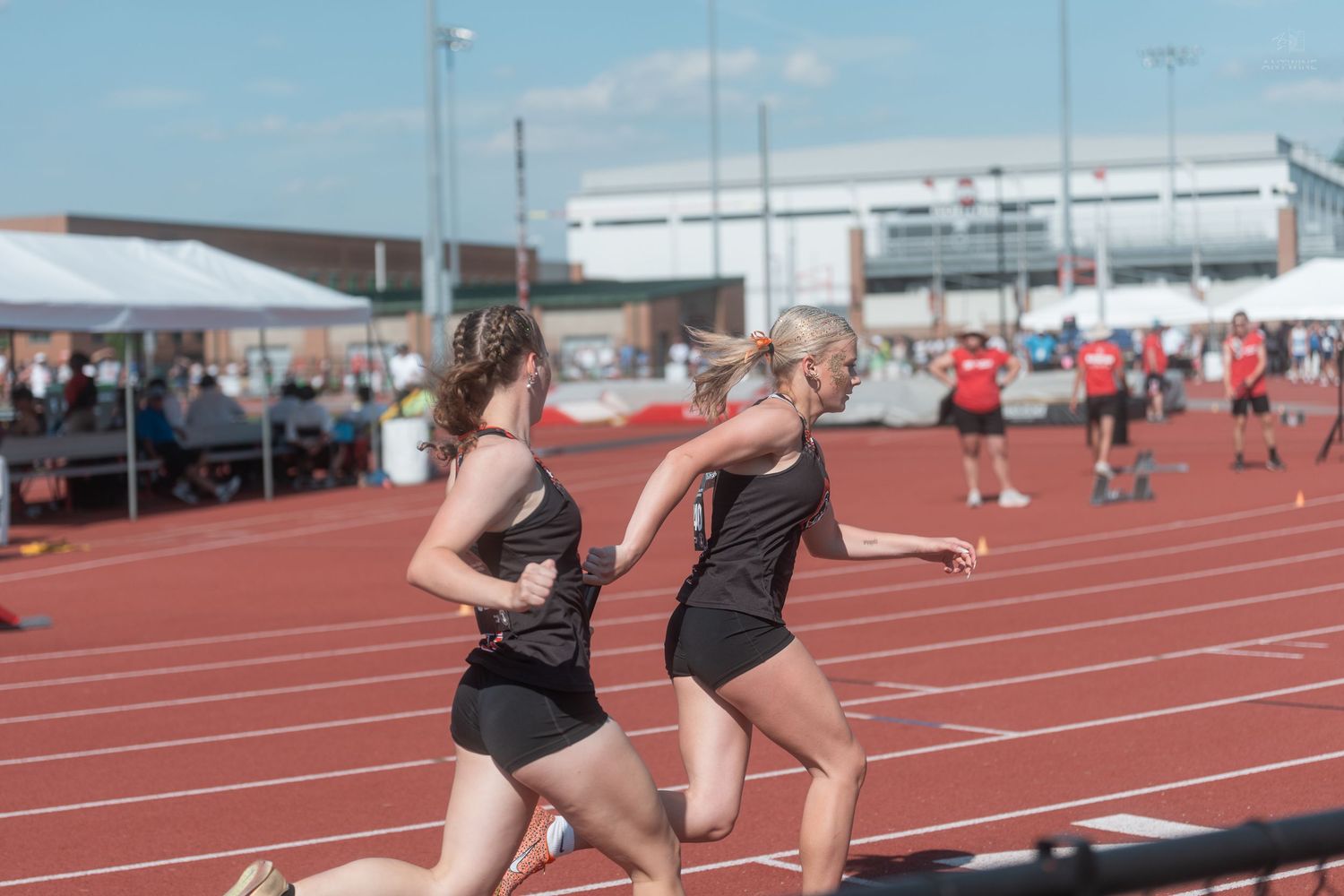 Two female athletes in black uniforms sprint side by side on a red track during a relay race.