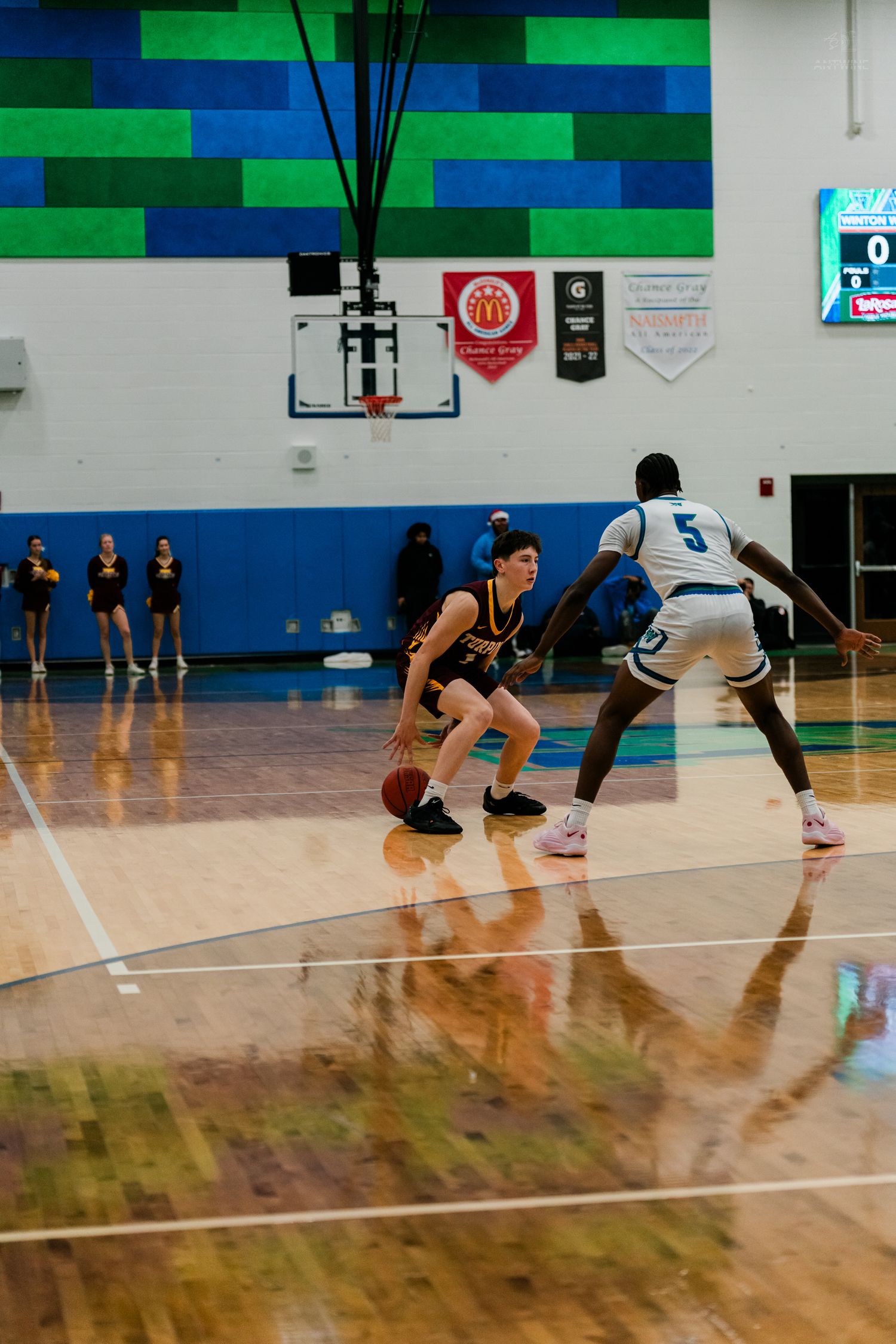 A player in black dribbles the ball while being defended by #5 in white during a high school basketball game.