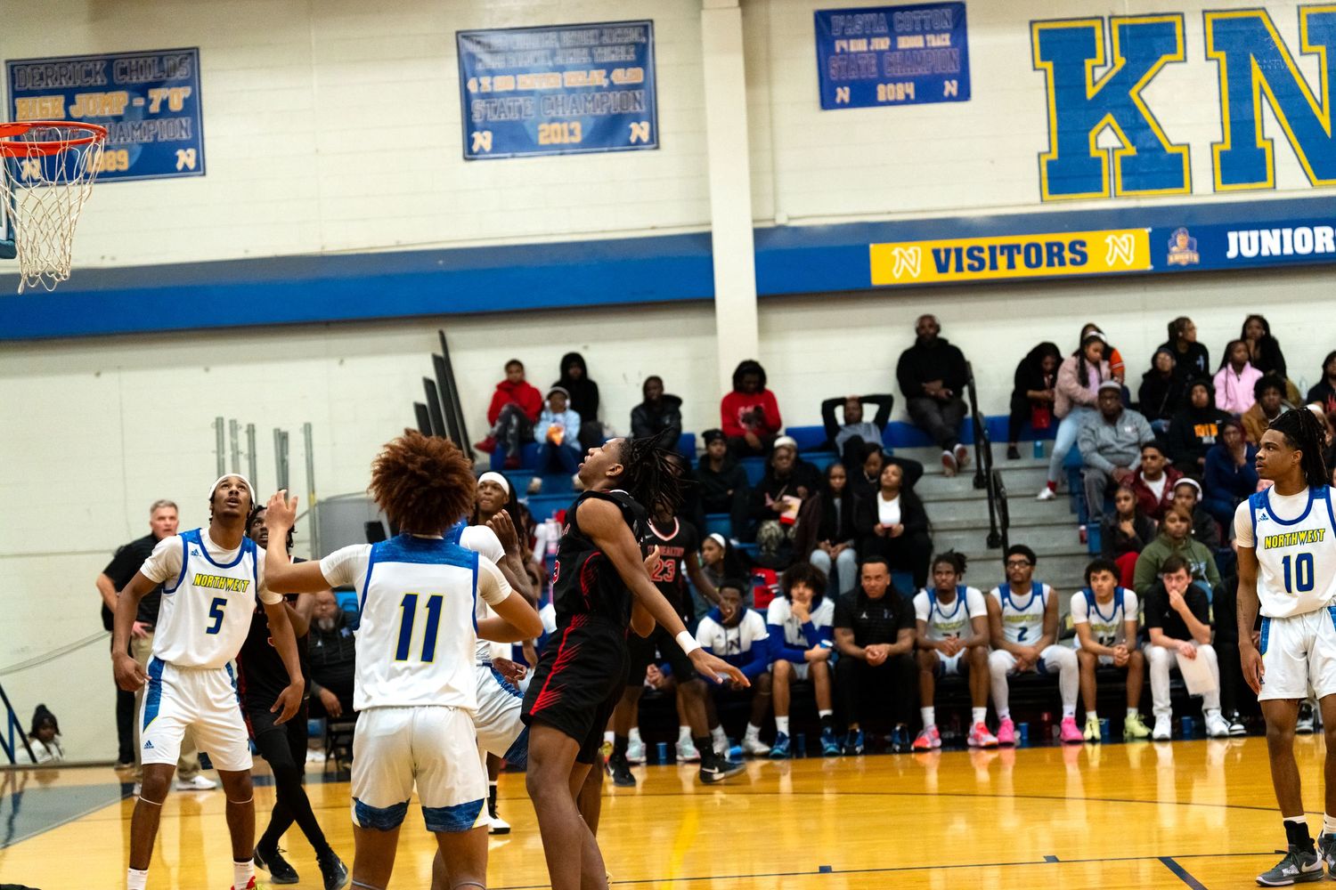 High school basketball game with players in blue and white uniforms defending against opponents in red and black.