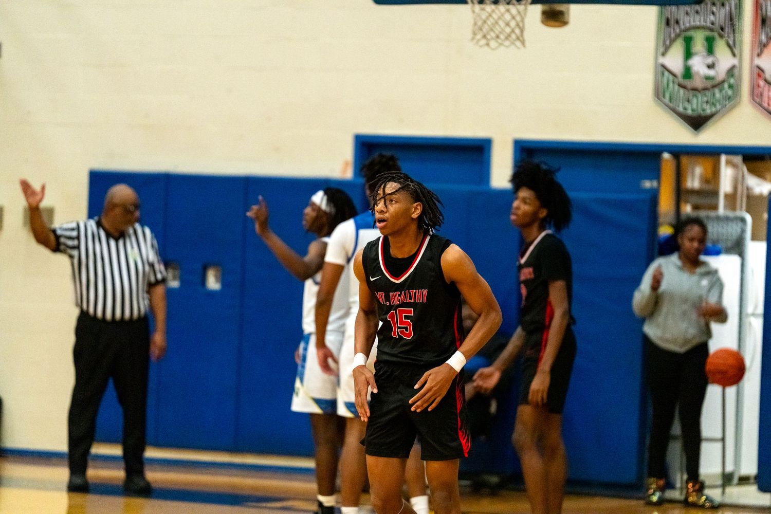 High school basketball player in red number 5 jersey stands confidently on the court near a referee.