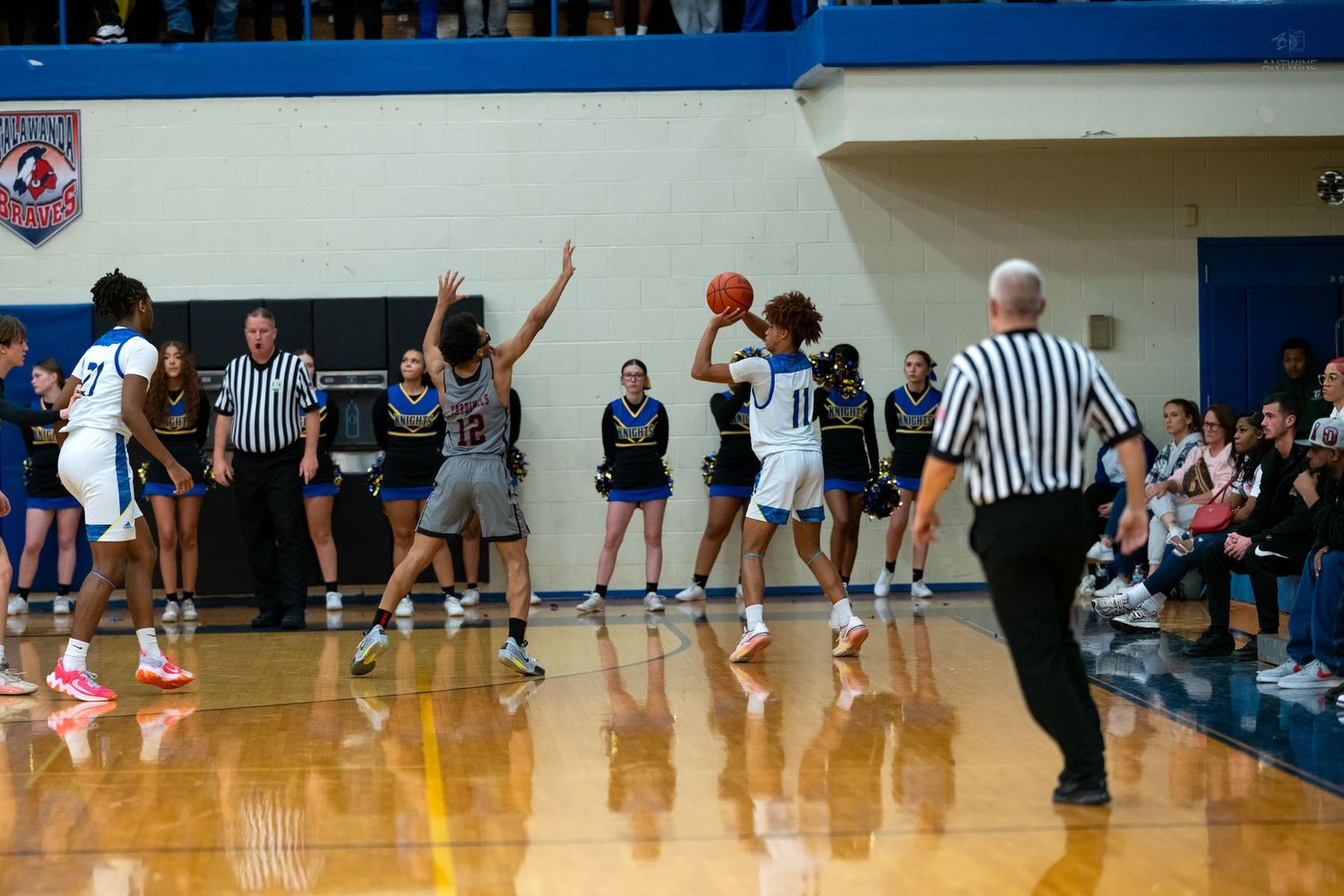 High school basketball player shoots jump shot while teammates and referee watch during indoor game.