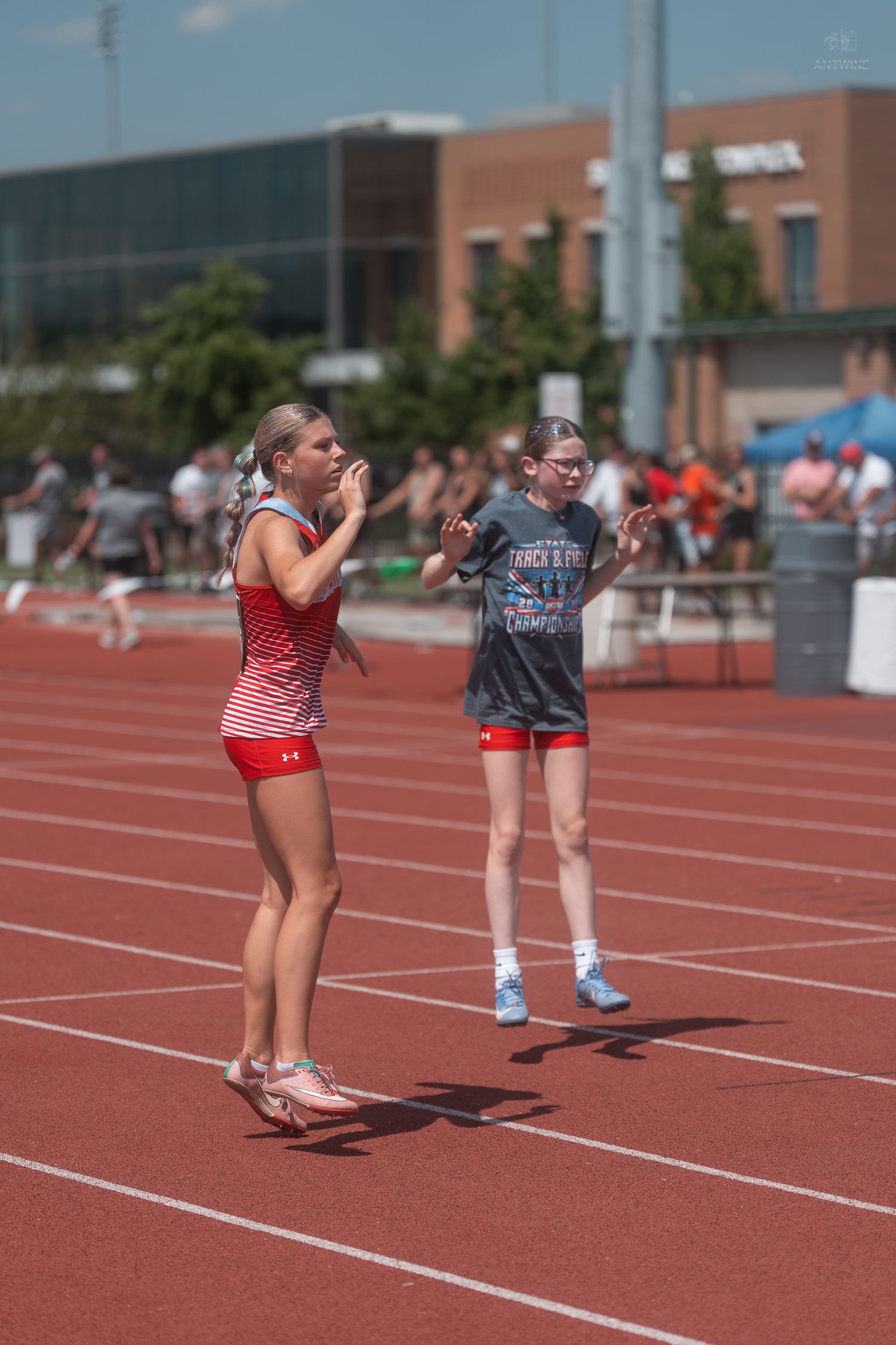 Two female track athletes in red and gray uniforms stand on red track watching competition at outdoor meet.