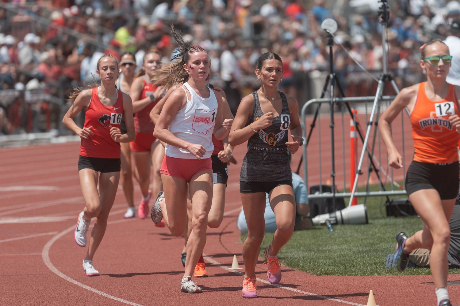 Female runners compete in a middle distance race at a sunny outdoor high school track and field championship meet.