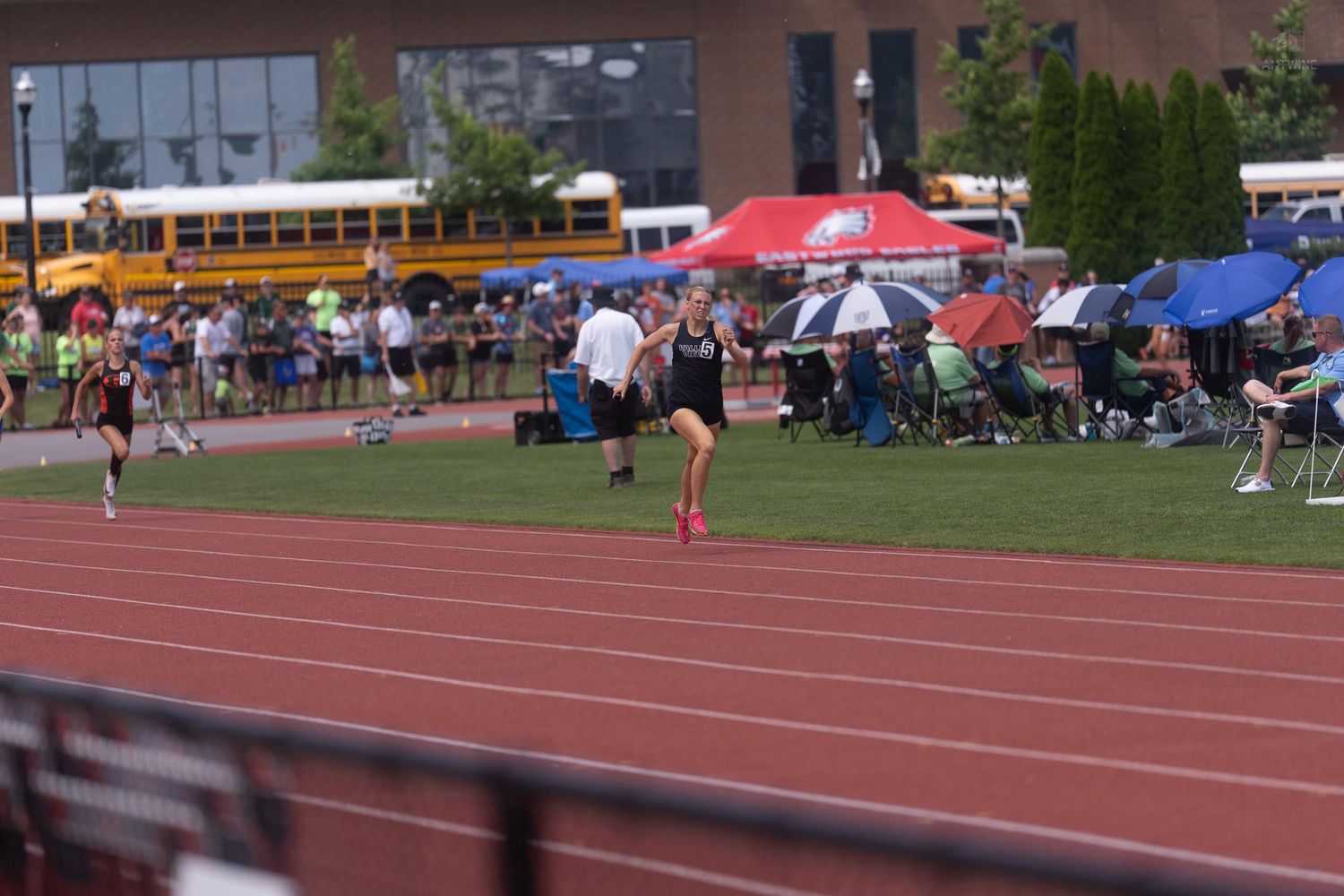 Athletes race on a red track at an outdoor track meet with school buses parked in the background.