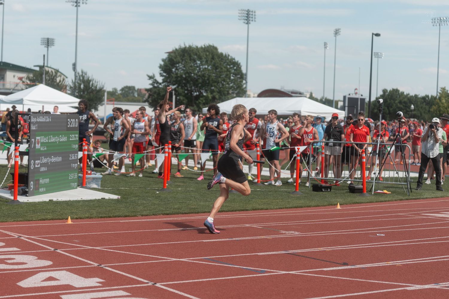 Runner competes on red track at outdoor track meet with large crowd of spectators lining the course.