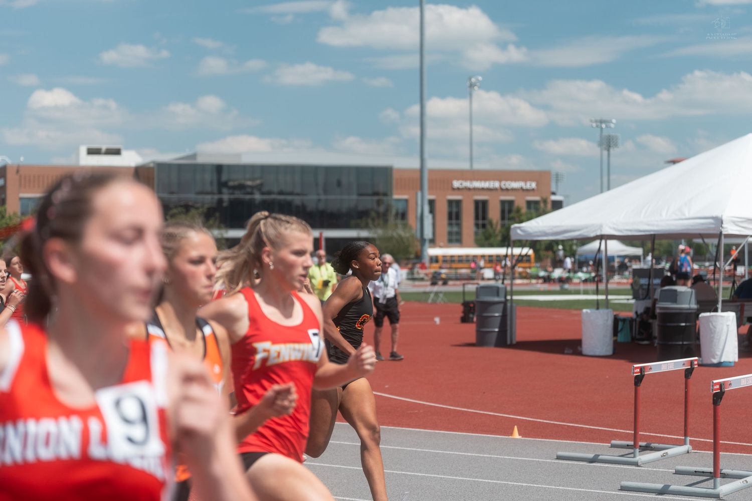 Female athletes competing in an outdoor track race with a red track and modern sports complex in the background.