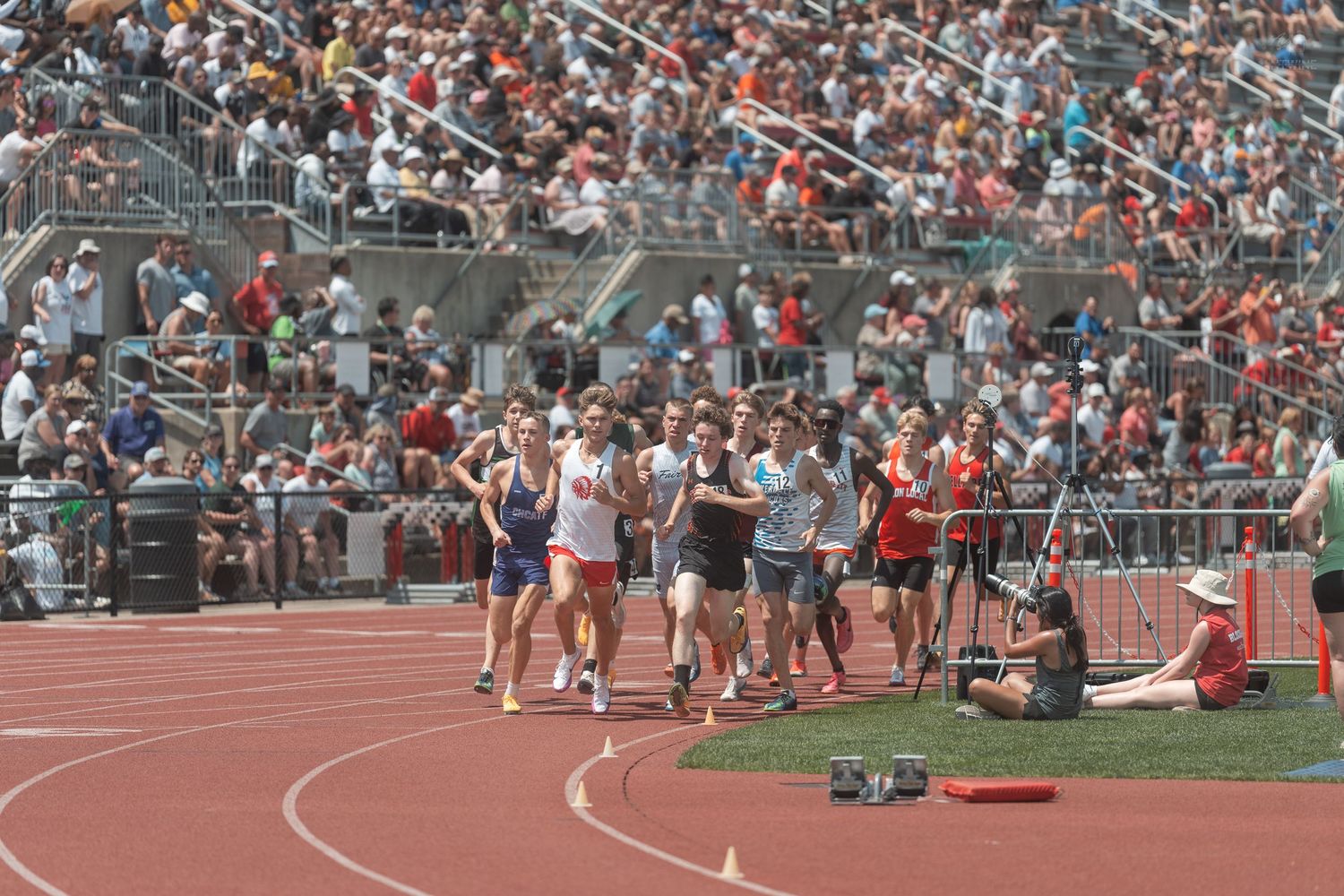 Competitive distance runners race on a red track at a high school track meet with a packed stadium crowd.
