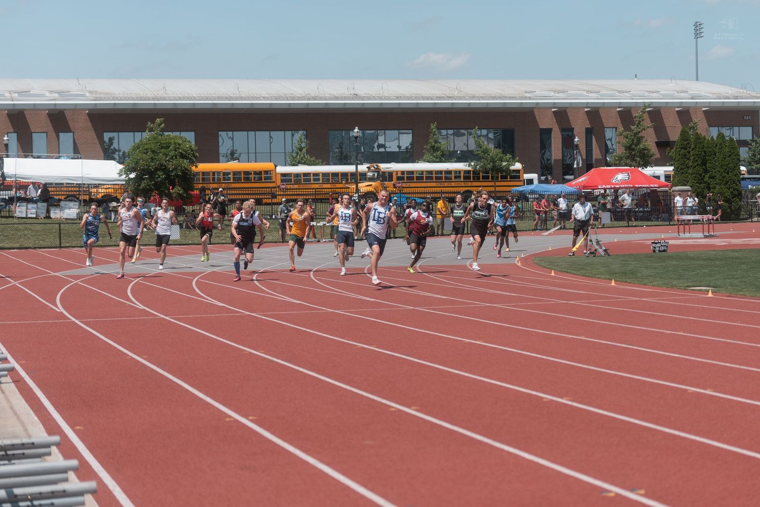 Runners race down the straightaway of a red outdoor track during a competitive high school track and field meet.