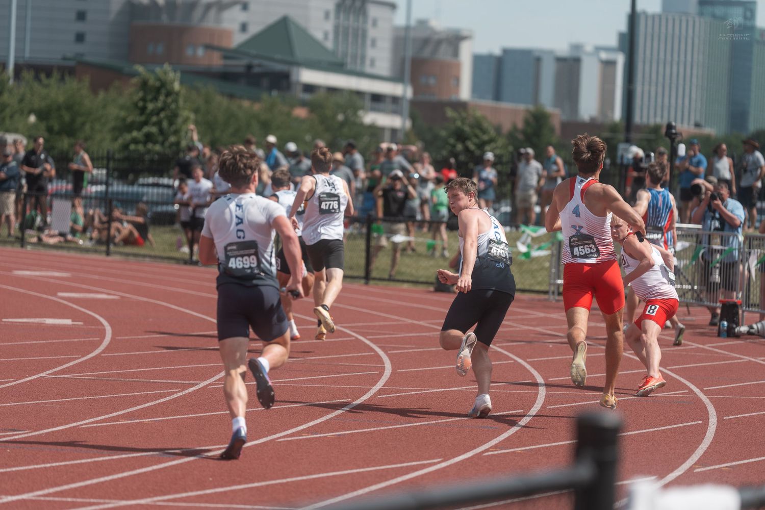 Multiple male runners compete in outdoor track relay race on red track in sunny conditions.