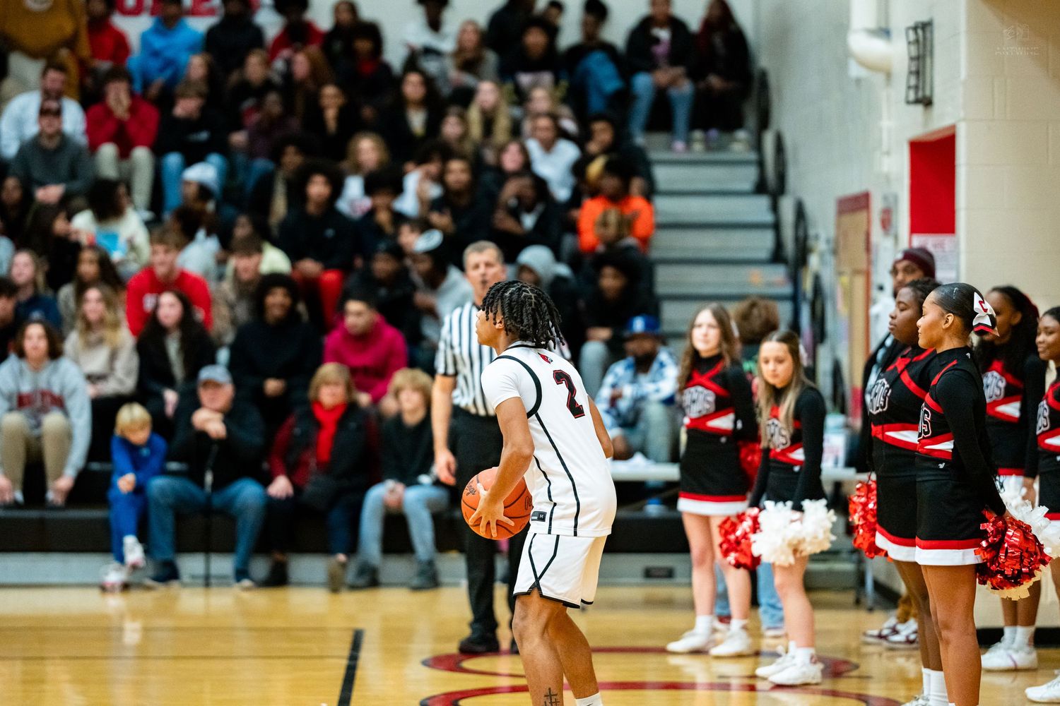 Basketball player in white uniform holds ball near cheerleaders and packed crowd during high school game.