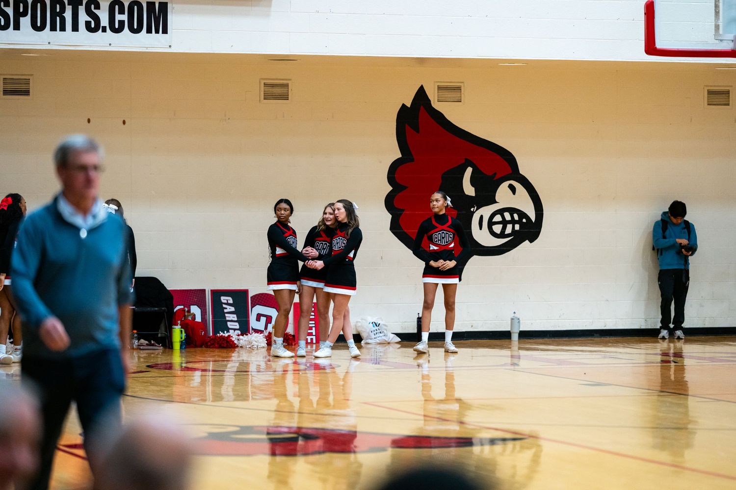 High school cheerleaders stand near a red Cardinal mascot logo painted on a gymnasium wall.
