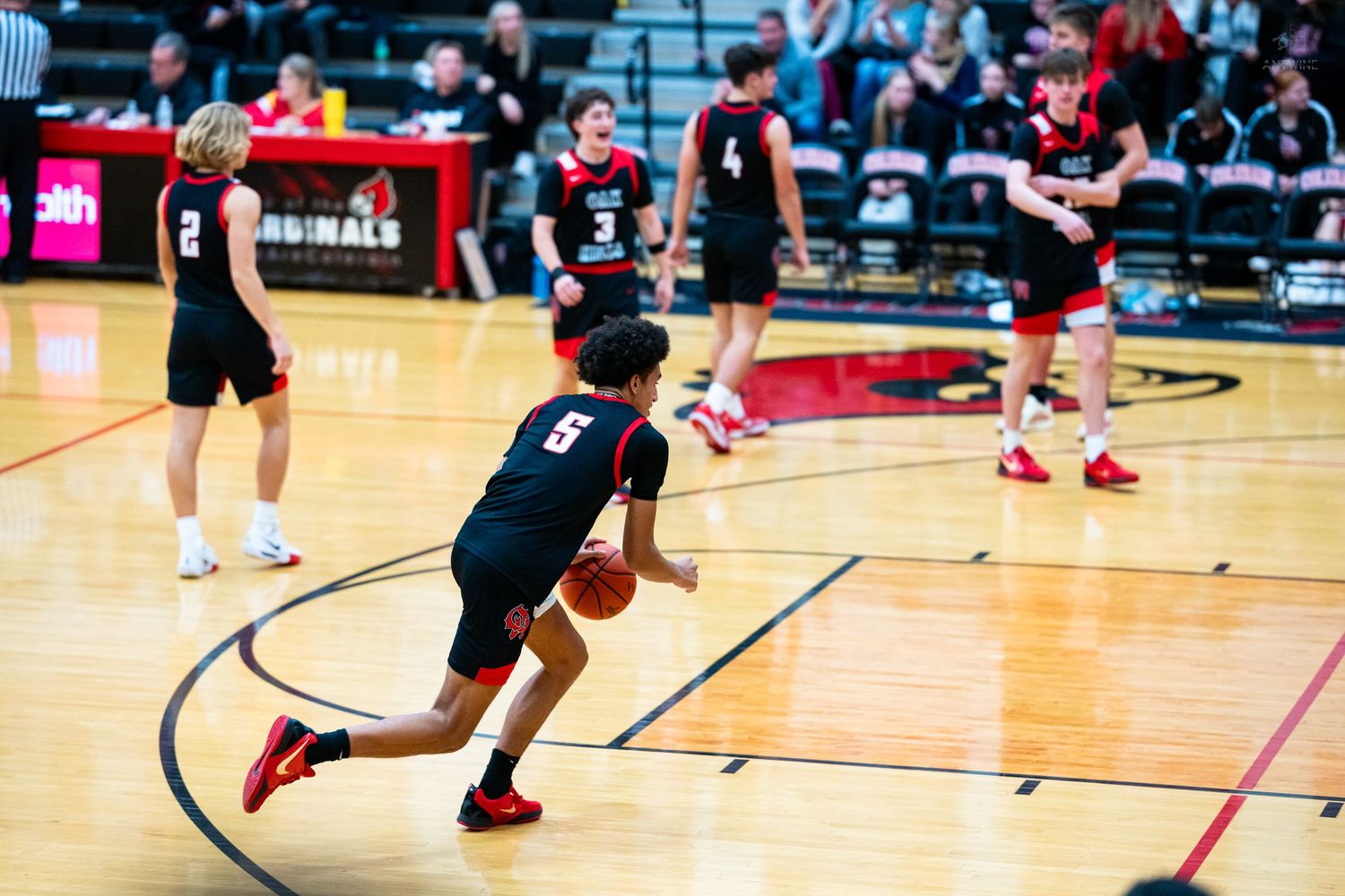 High school basketball player number 6 dribbles upcourt during a game in a red Cardinals gymnasium.
