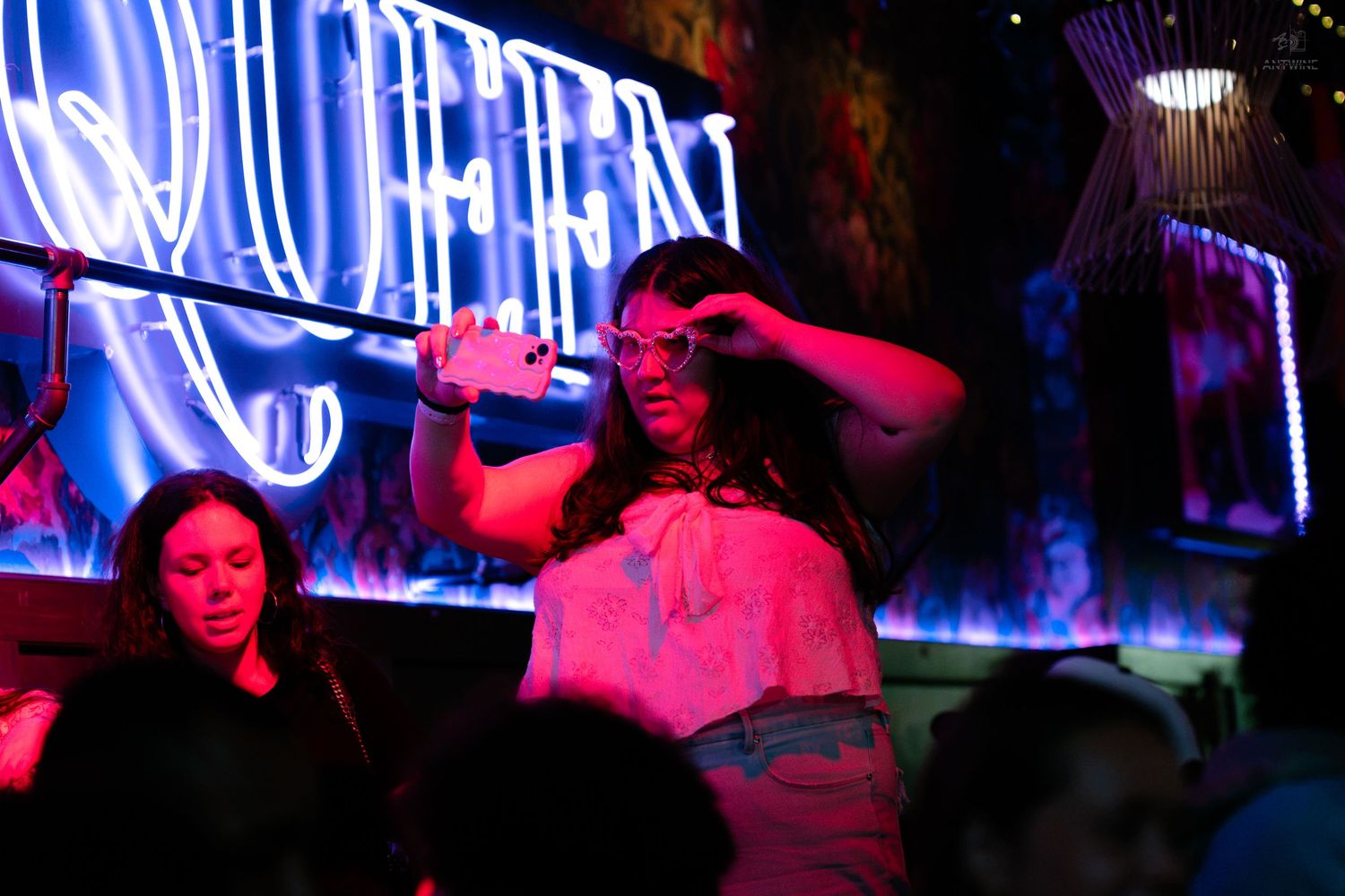 Woman posing with a drink under a neon QUEER sign at a vibrant nightclub.