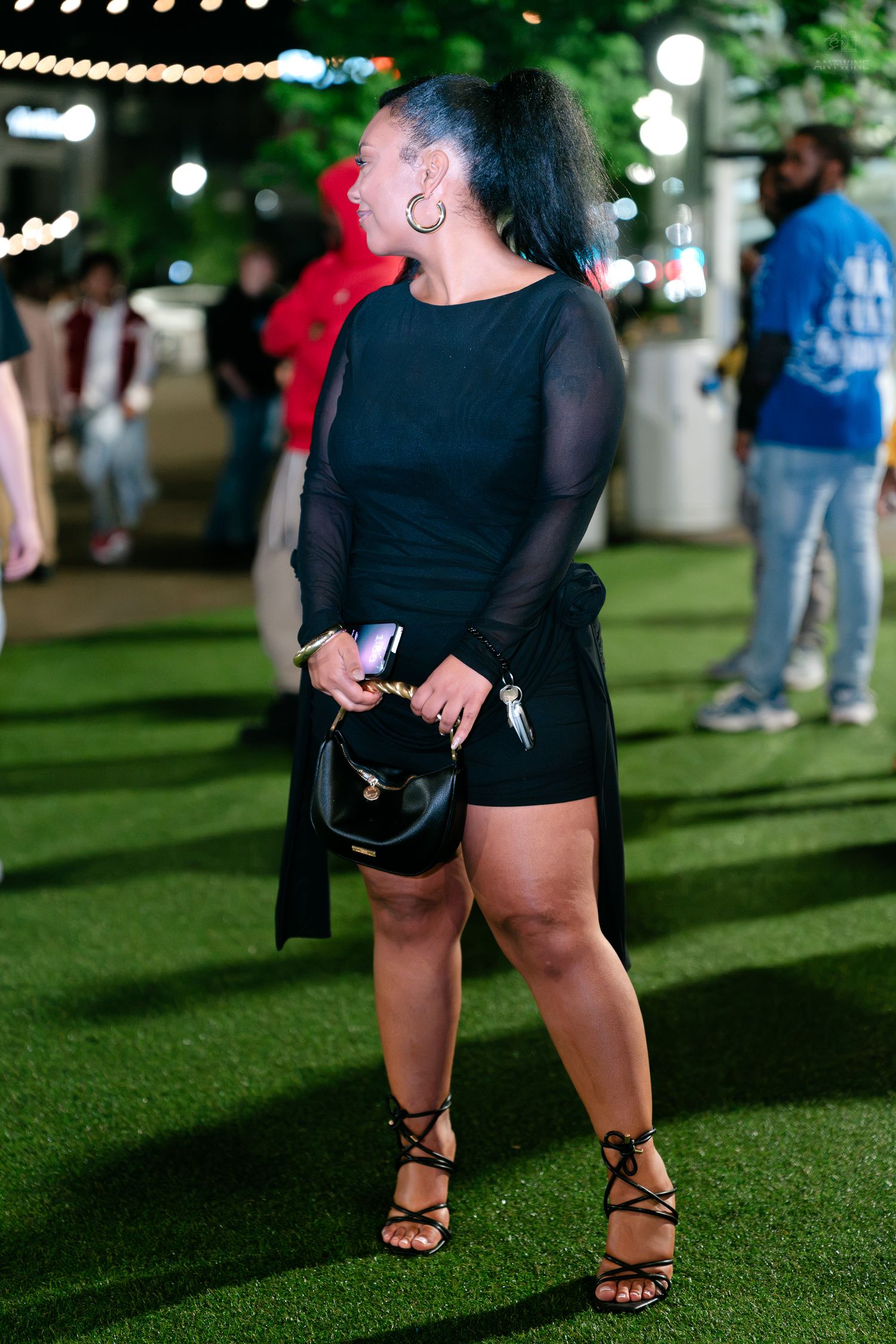 Woman in black mini dress with mesh sleeves holding handbag at outdoor night event.