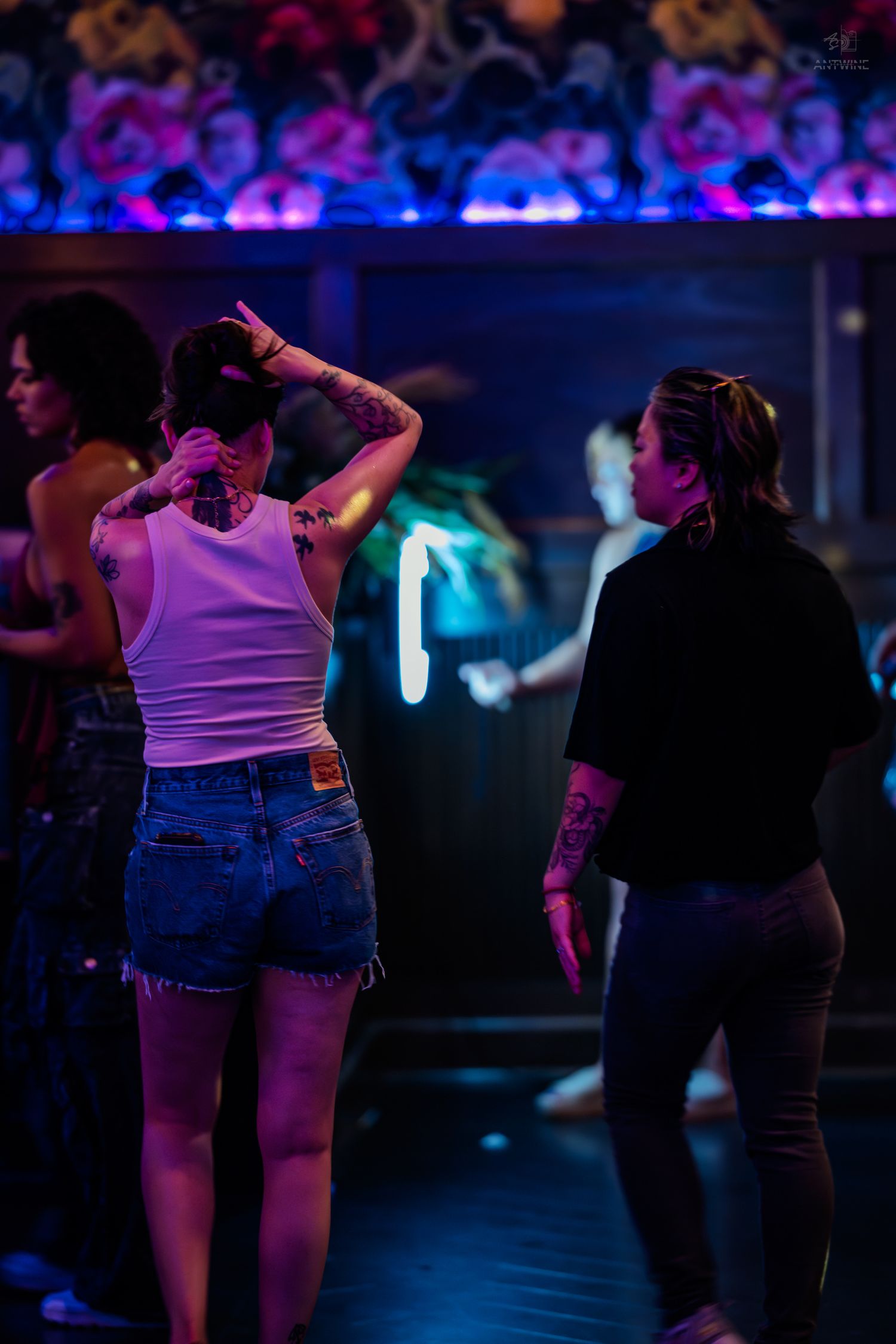 Two tattooed women dance under neon purple lights in a nightclub with floral decor.