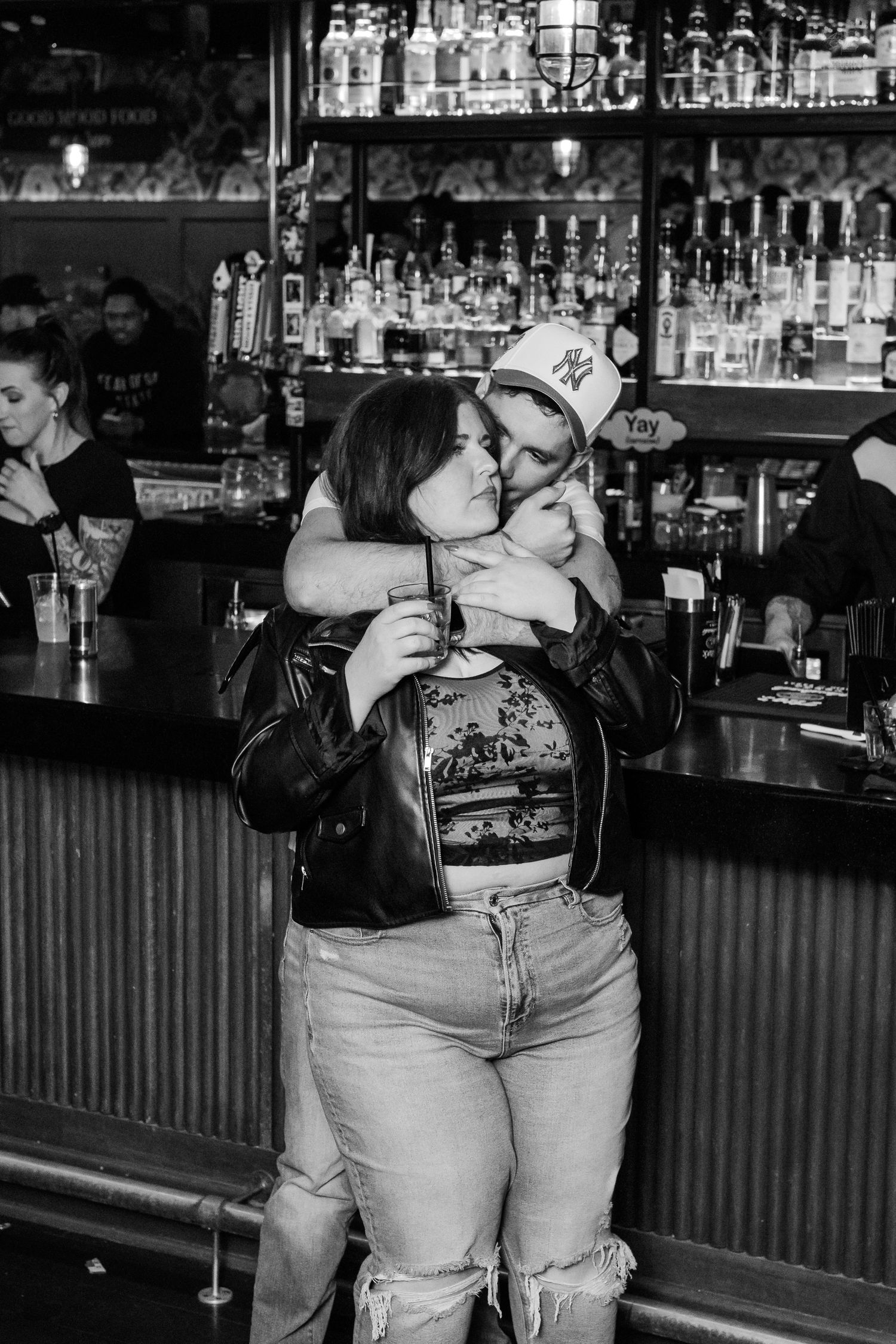 A couple embraces and shares a kiss in front of a well-stocked bar in a nightclub setting.