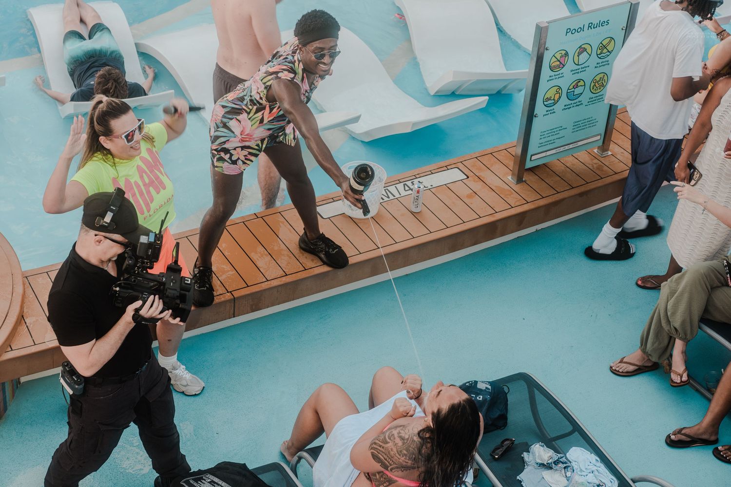 Videographer filming cruise ship guests relaxing and playing near a bright blue pool deck area.