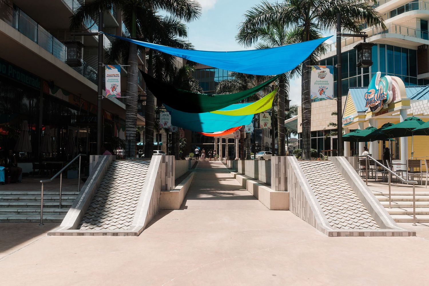 Colorful shade sails hang over a modern outdoor shopping plaza with palm trees and ramps in a tropical city.