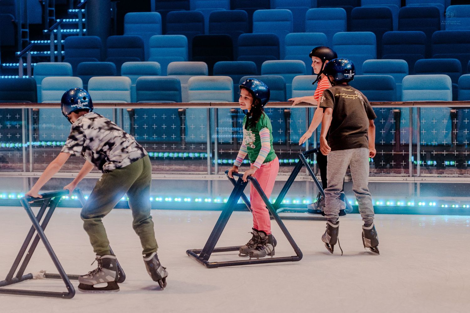 Kids wearing helmets practicing roller skating with metal support frames on a bright indoor cruise ship rink.