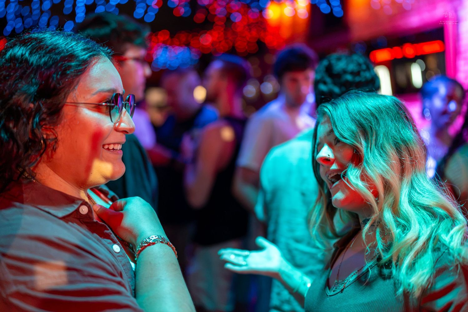 Two women smiling and chatting at a vibrant nightclub with colorful neon lights and crowd in background.