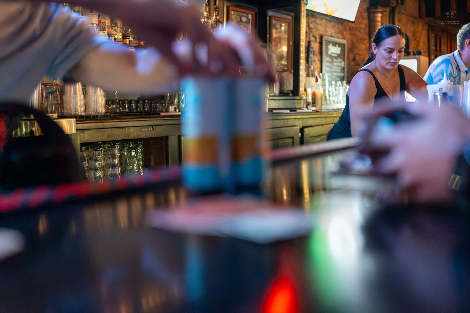 Bartender serving drinks at a dimly lit bar with blue cans on the counter and a woman in the background.