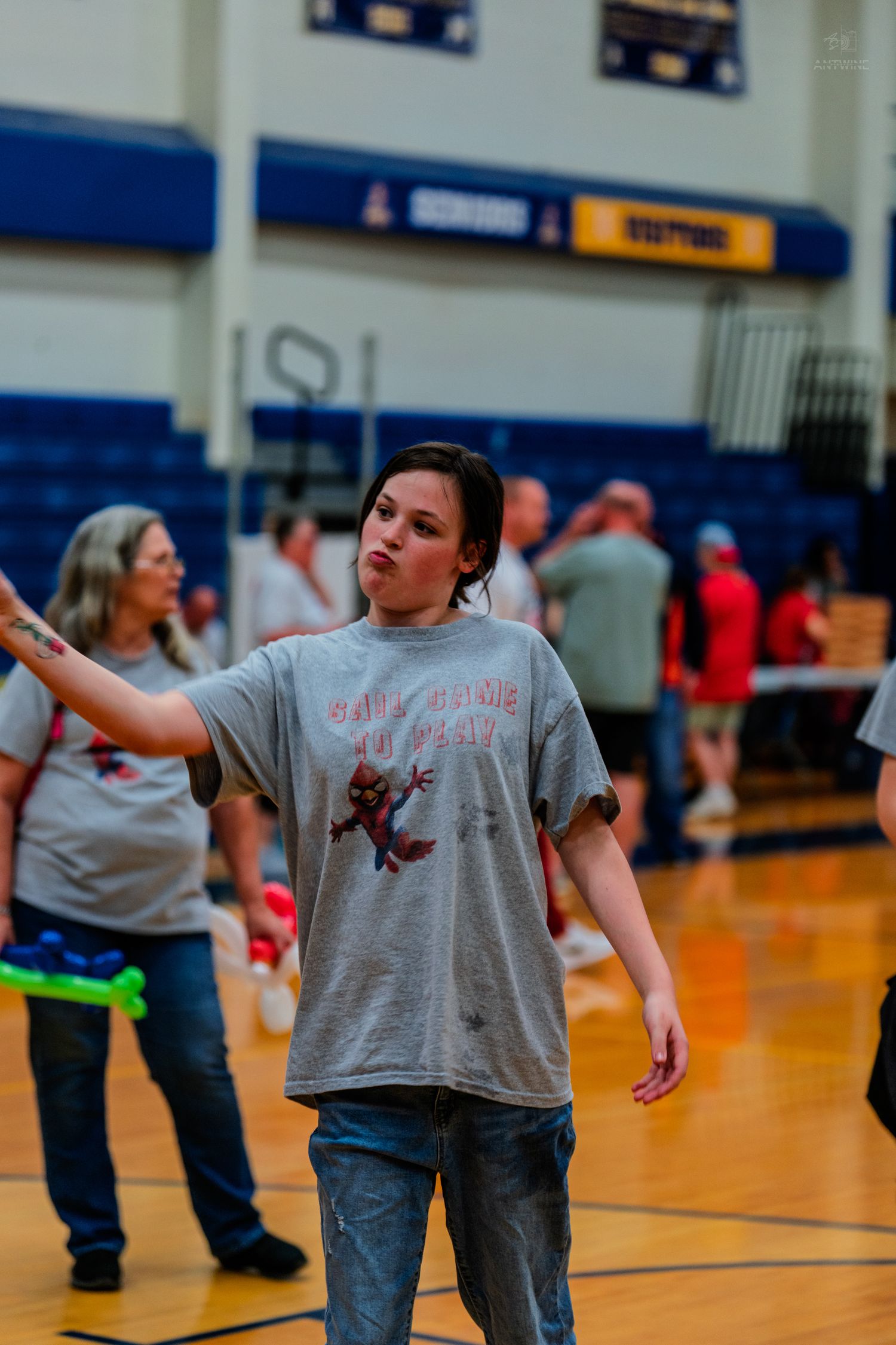 Young woman in gray graphic t-shirt gesturing on a gymnasium basketball court during a school event.