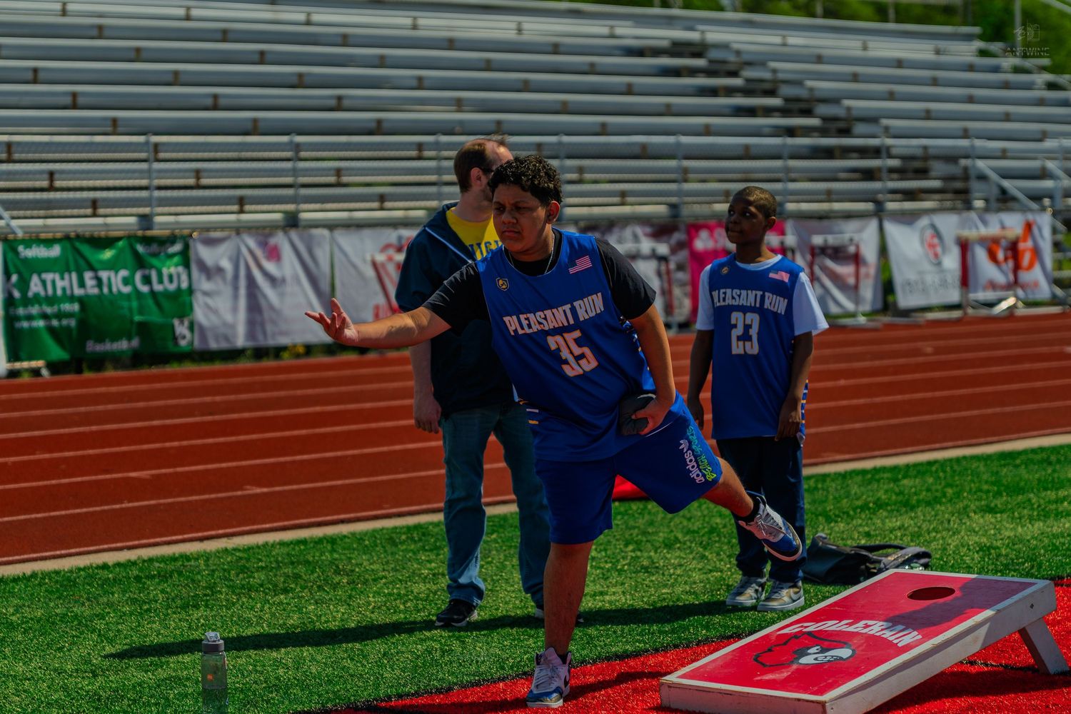 Pleasant Run team players compete in a cornhole game on a red track field at an outdoor athletic stadium.