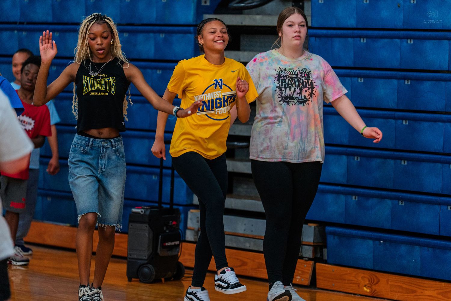 Three students in Knights school shirts dance and cheer together on a gymnasium floor during a school event.