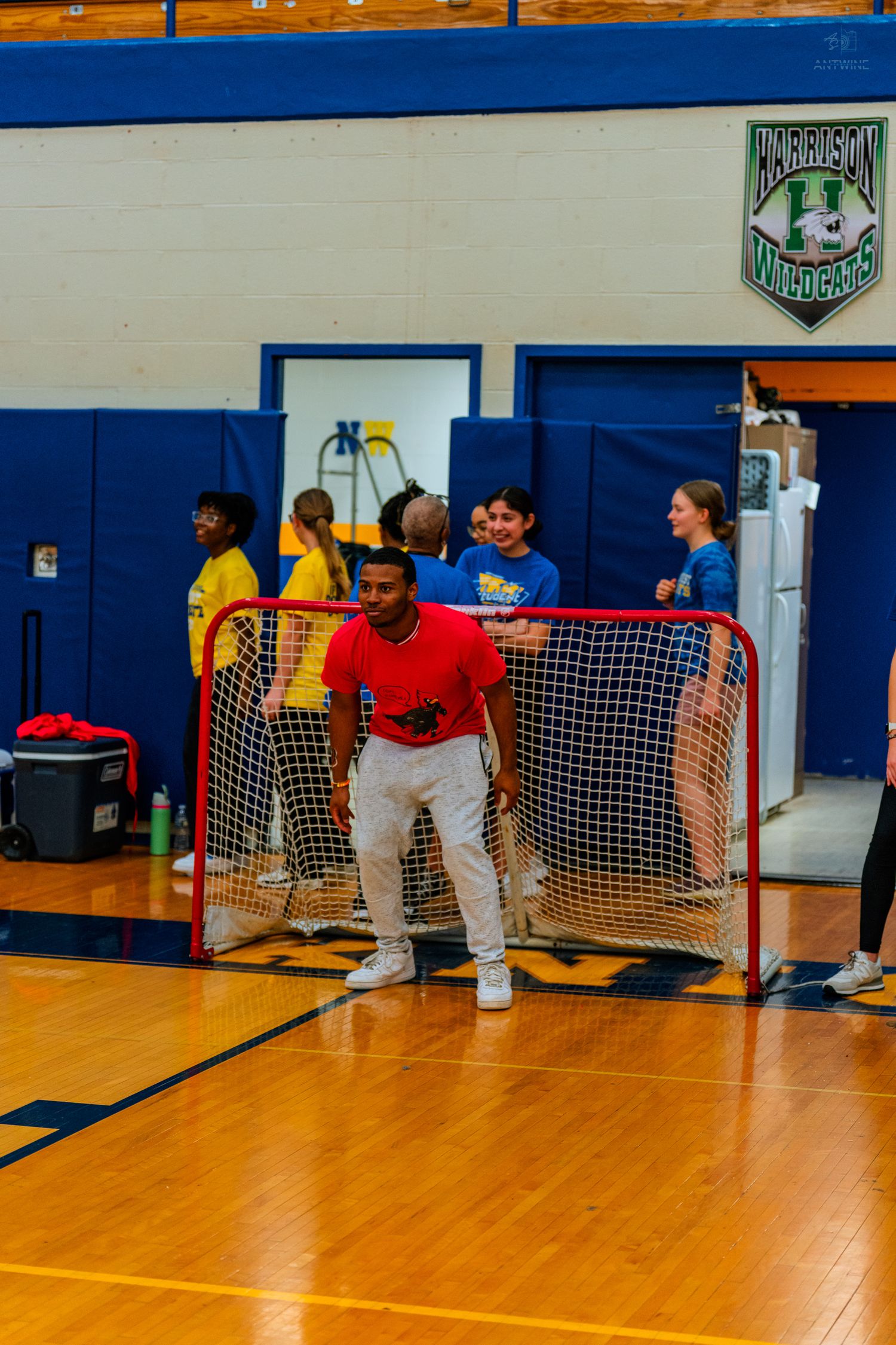 Student in red shirt plays goalie in indoor floor hockey game in school gymnasium with Wildcats logo on wall.