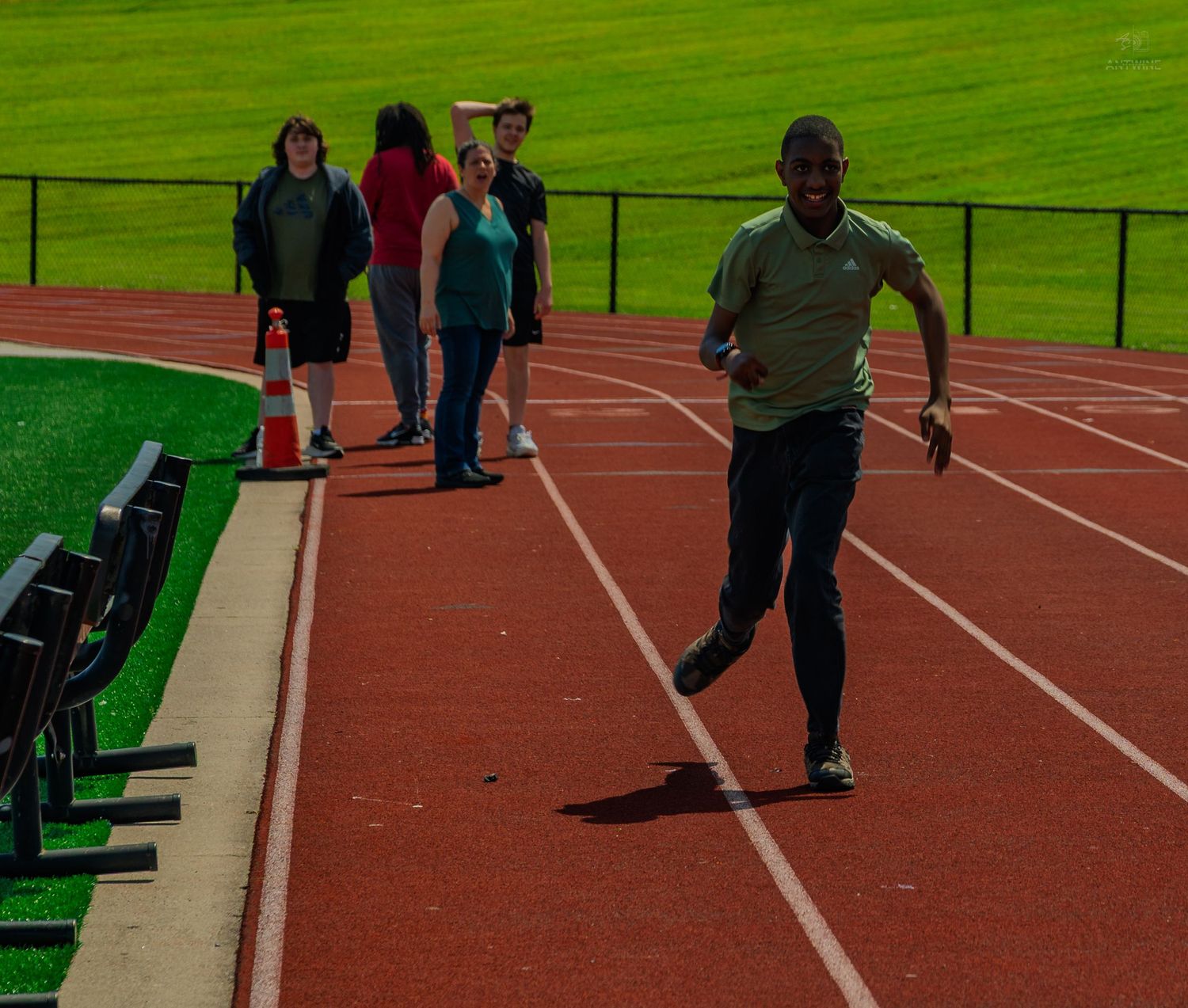 Athlete sprinting on a red running track while spectators watch from the sideline on a sunny day.
