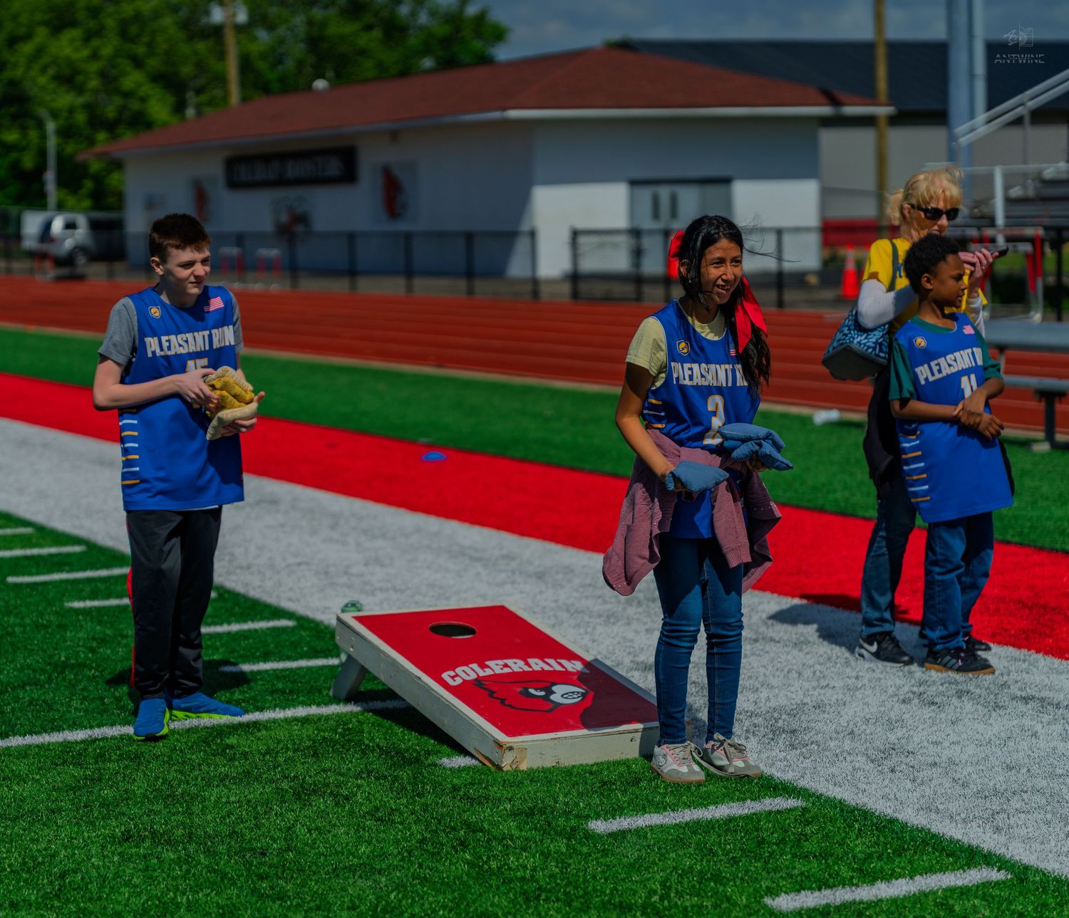 Students in Pleasants blue jerseys play cornhole on a red and green athletic field during a school sports event.
