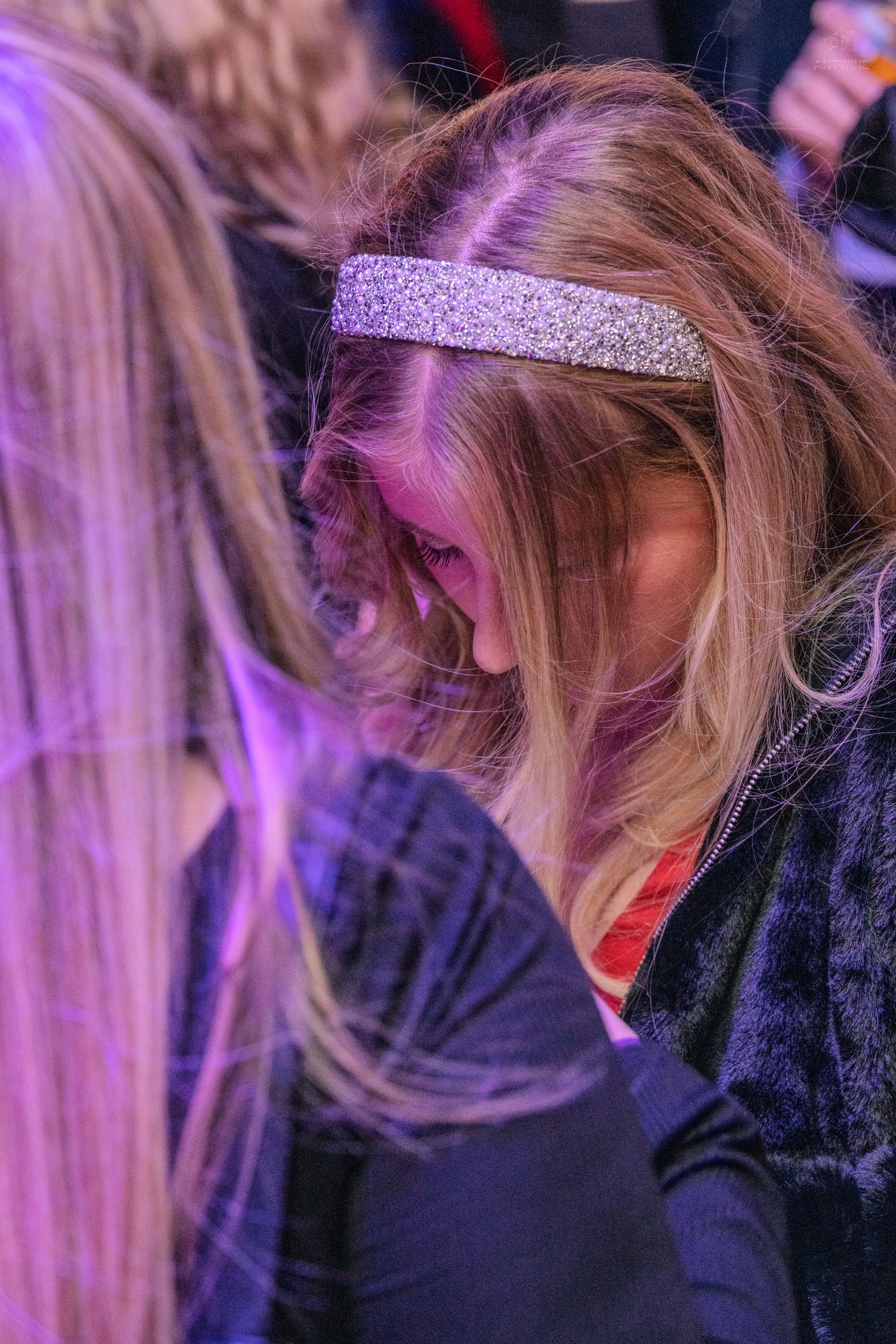 Blonde woman with glitter headband laughing at a crowded nightclub event.