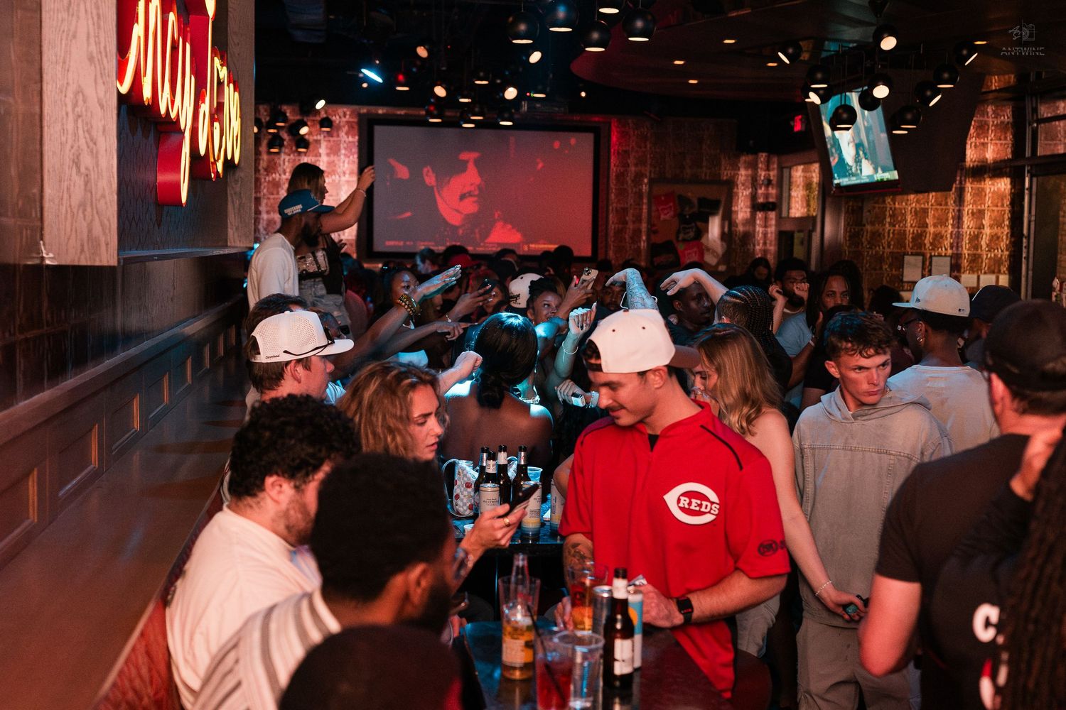 Lively sports bar crowd in Cincinnati Reds apparel watches game on large projection screen.