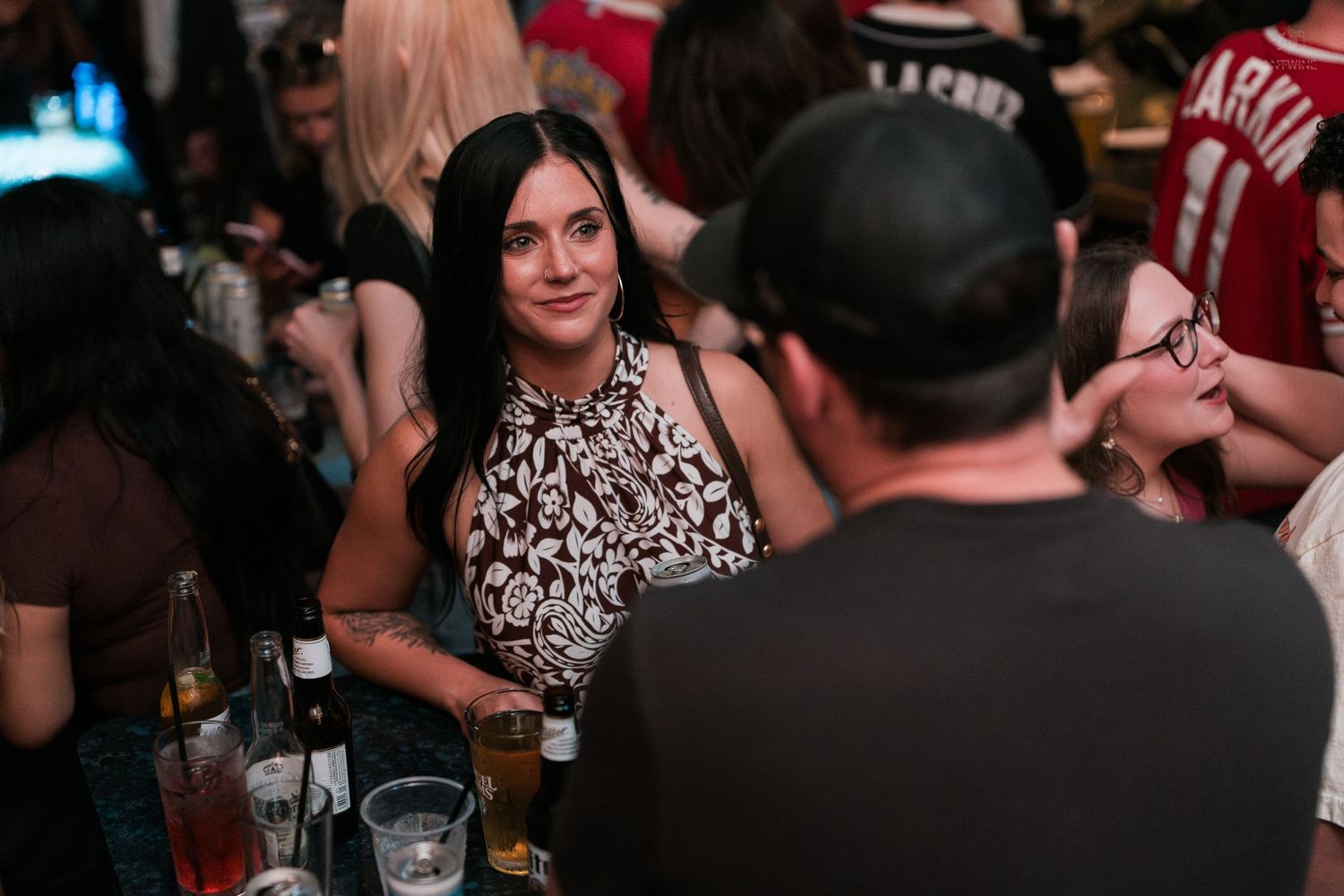 A dark-haired woman in a floral top smiles at a man while holding a drink at a busy bar.