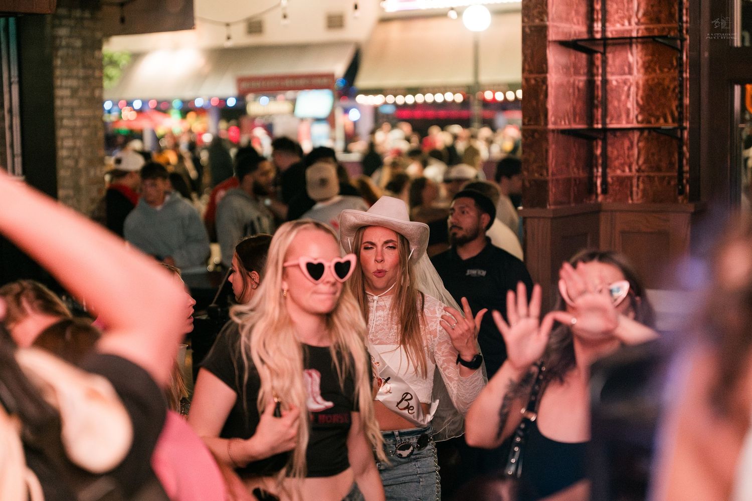 Two blonde women posing and dancing in a busy bar, one wearing heart-shaped sunglasses and a white cowboy hat.