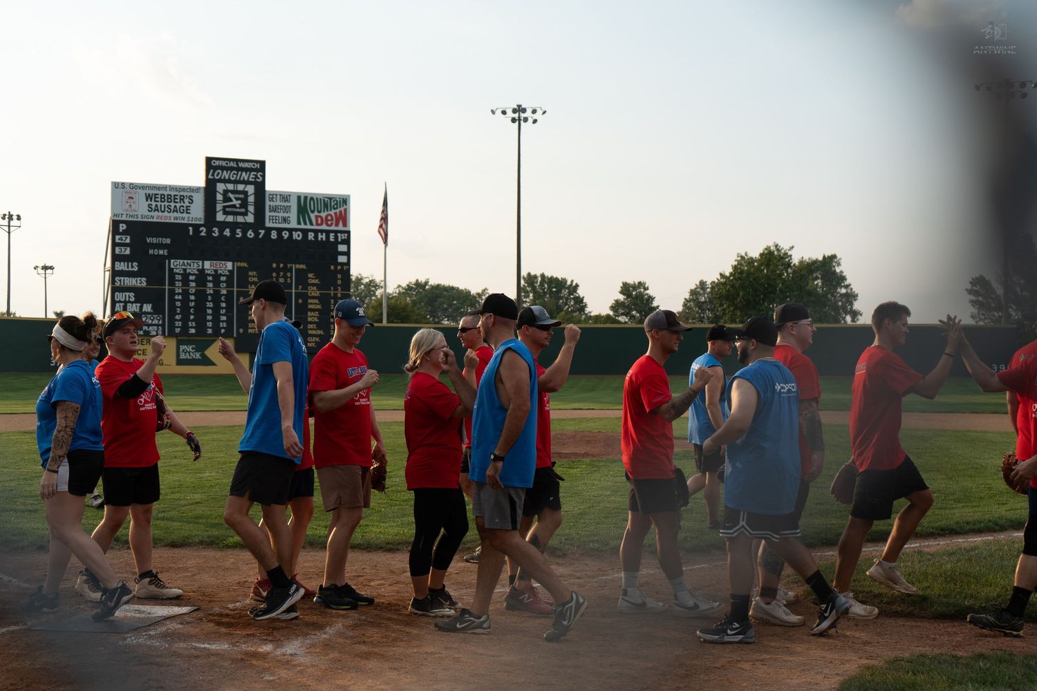 Teams shake hands on a baseball field after a game, with a scoreboard visible in the background at dusk.