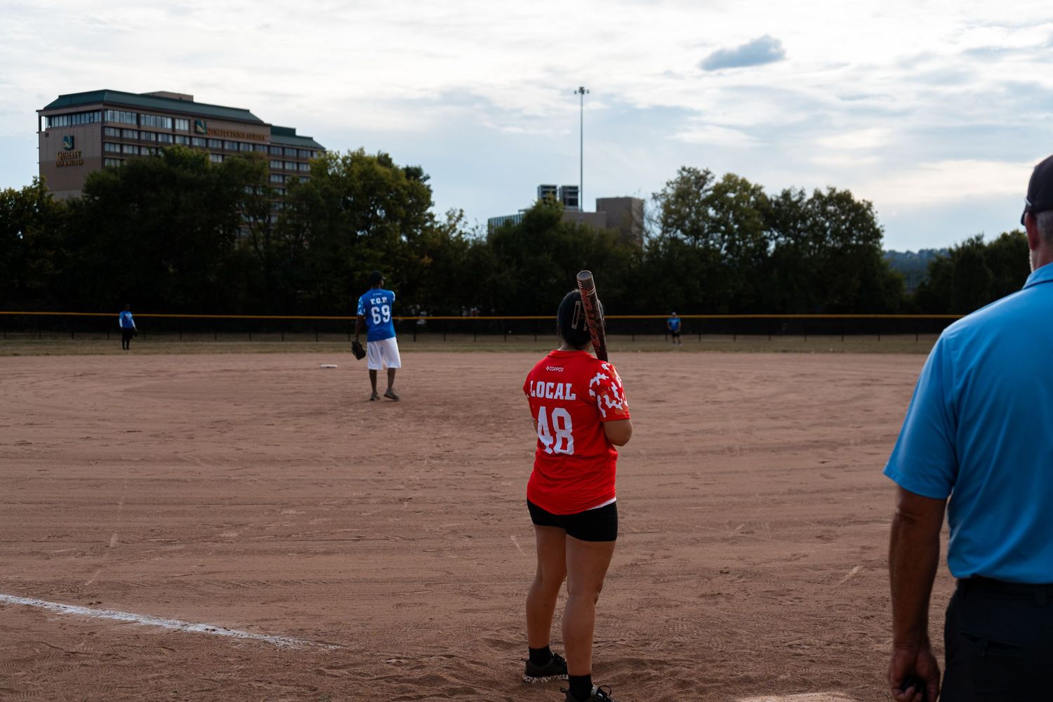 Woman in red jersey #49 playing softball on a dirt field with players and urban buildings in the background.