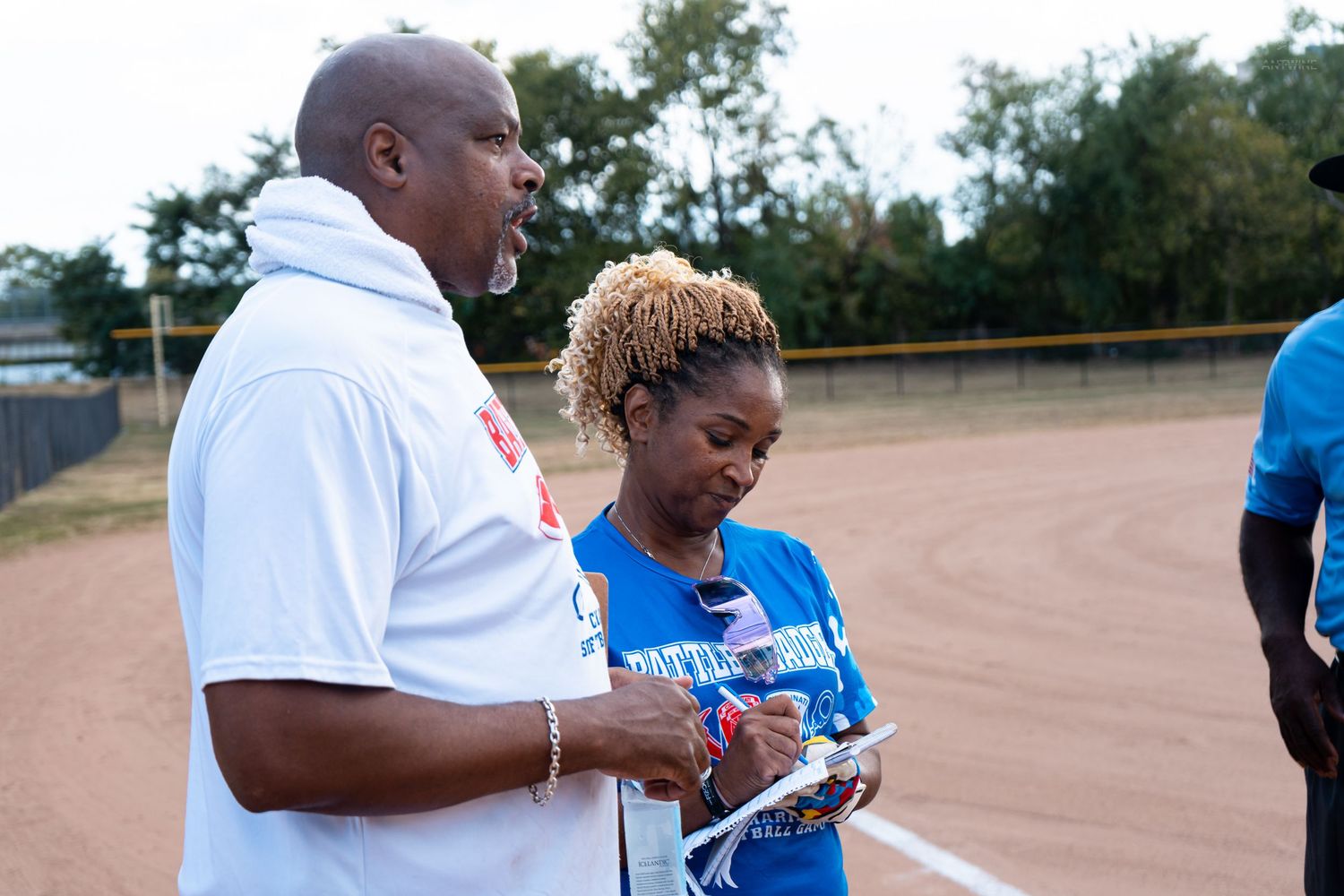 Man in white shirt and woman in blue jersey review paperwork together on a softball field.