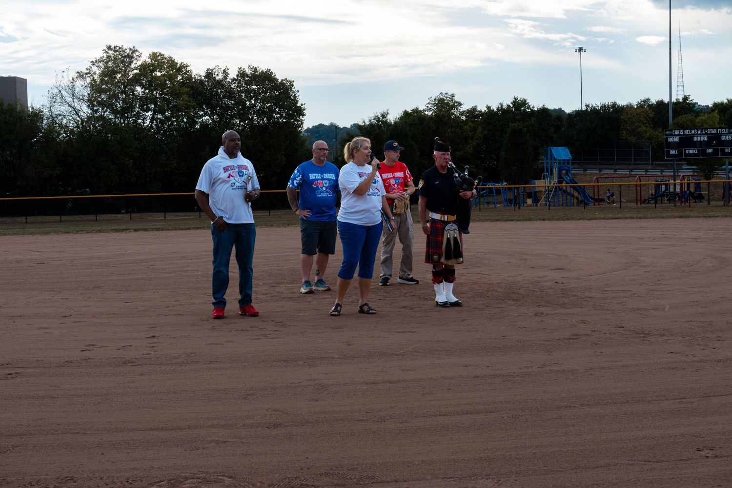 Group of five people with a bagpiper in kilts gathered on a sandy softball diamond at twilight.