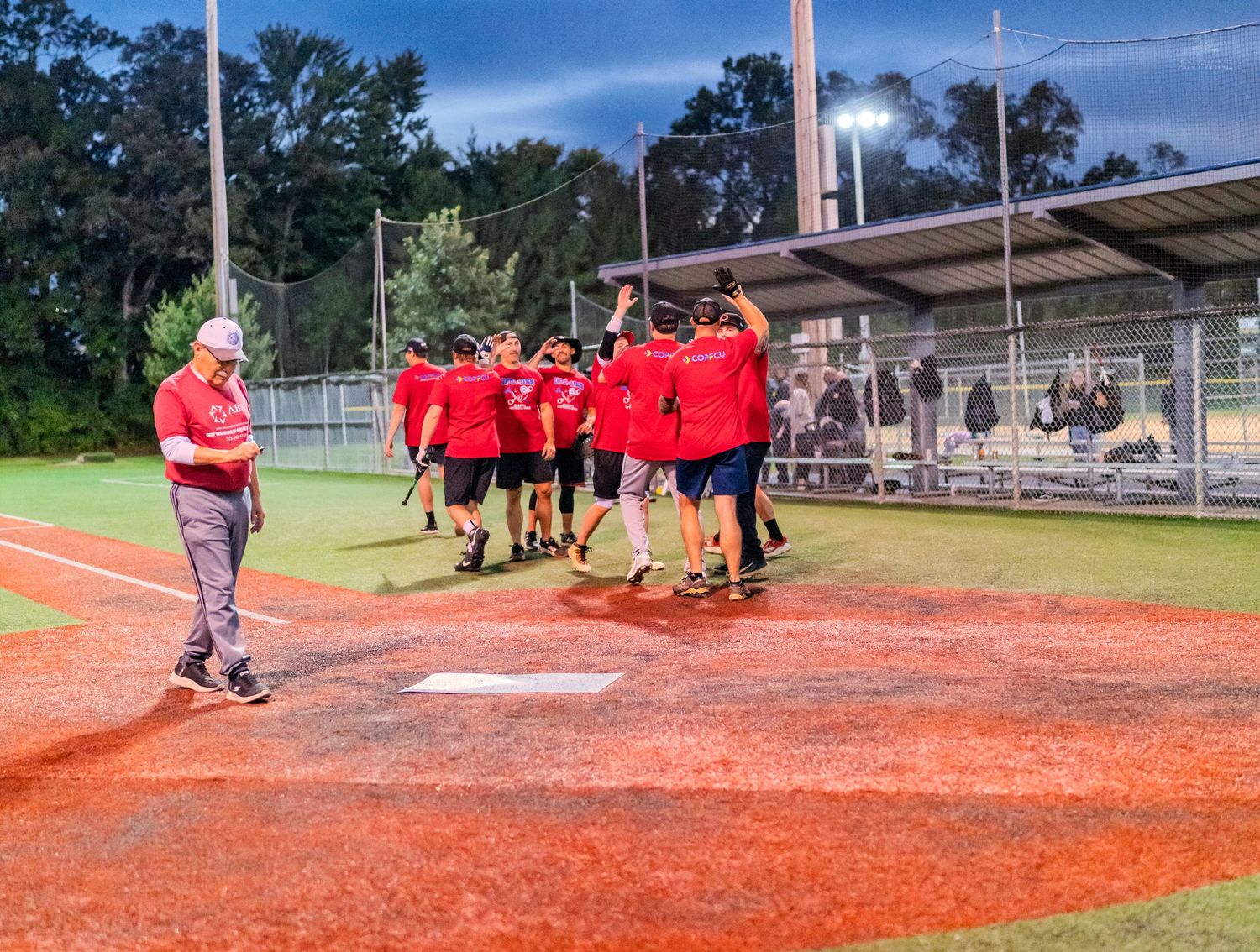 Red-shirted softball team celebrates with high-fives near home plate under stadium lights at dusk.