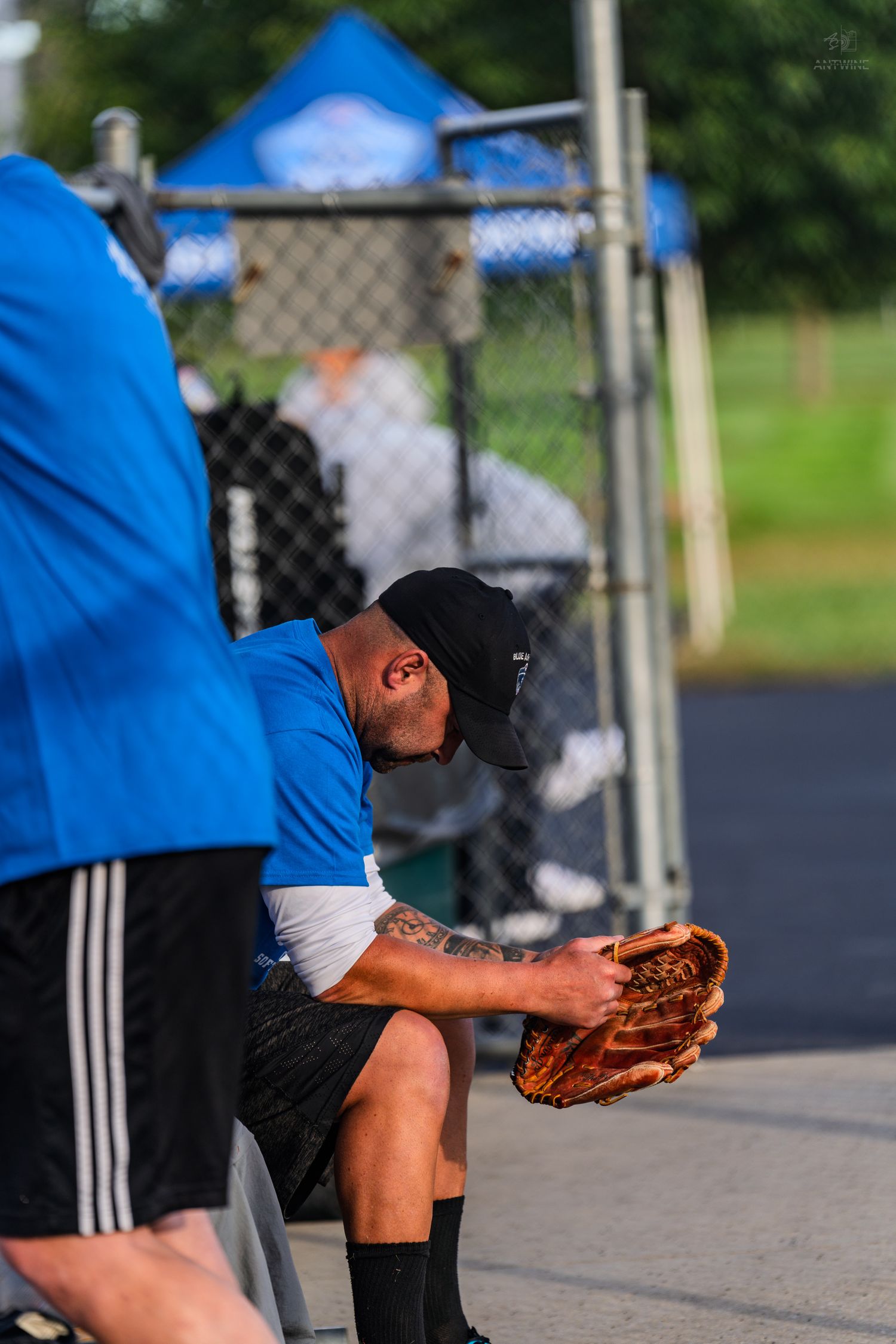 A baseball player in a black cap examines his worn leather glove while sitting in a dugout.