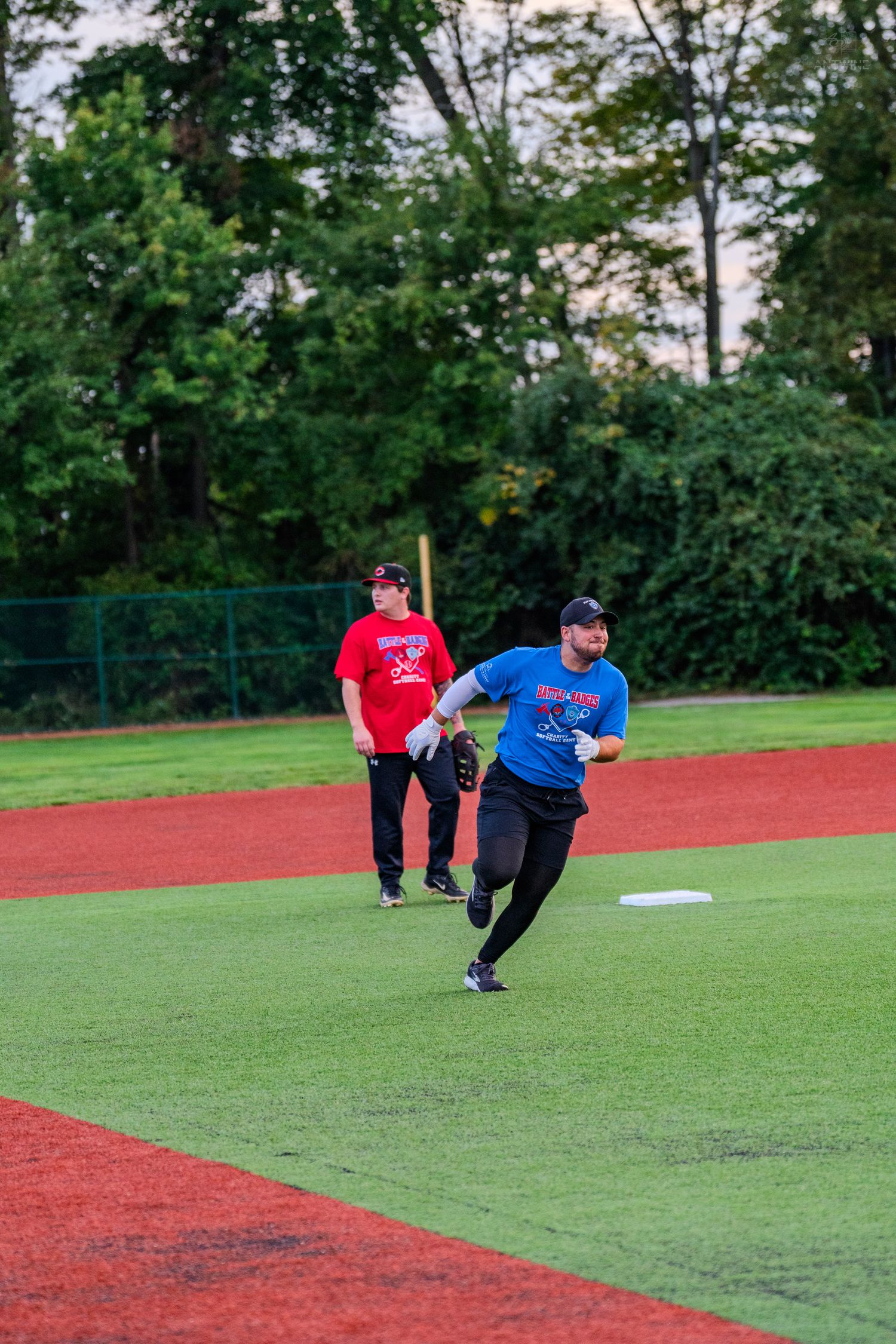 Two men on baseball field, one in blue shirt throwing ball while another in red watches nearby.