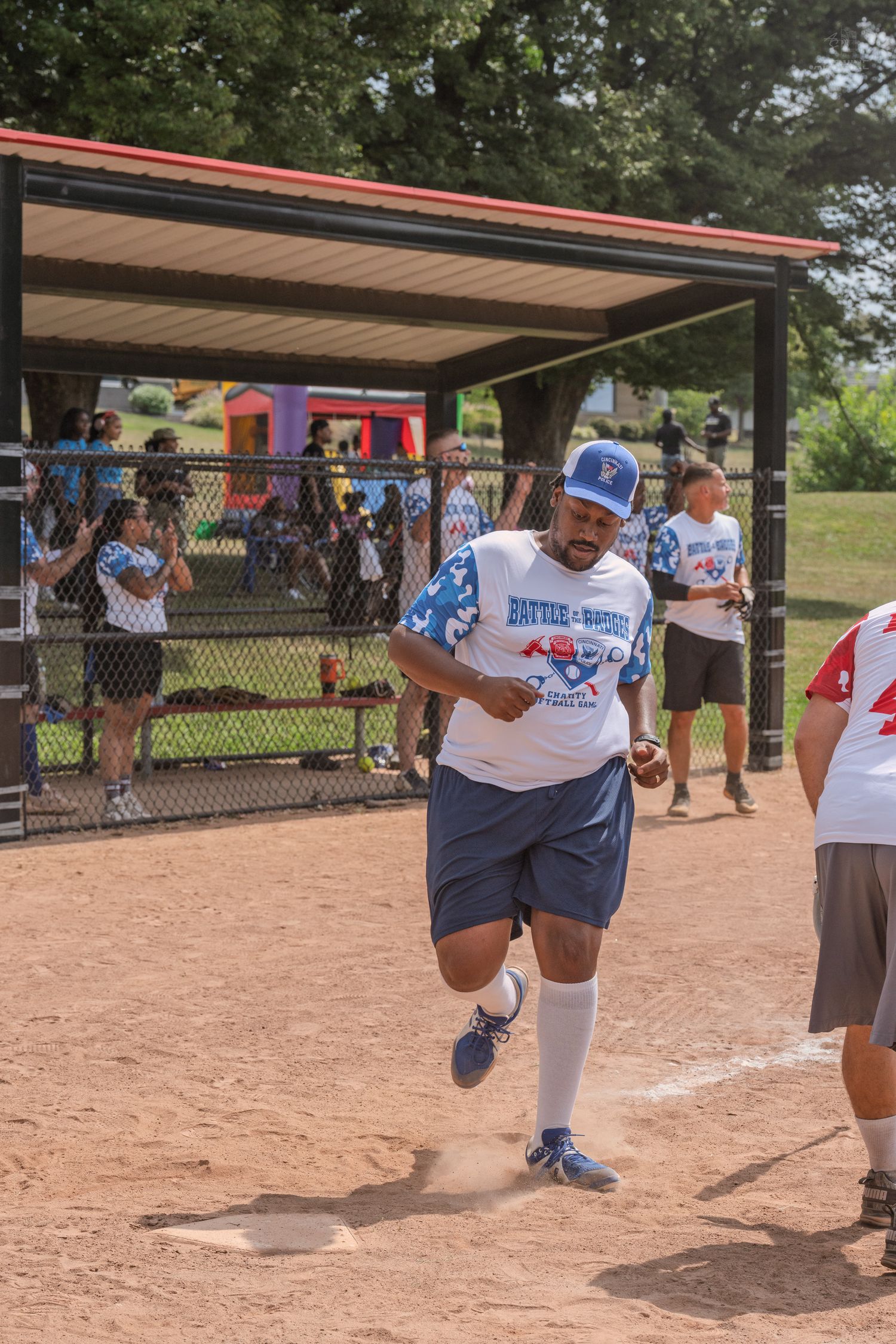 Smiling man in blue cap and charity softball jersey runs bases on dirt infield.