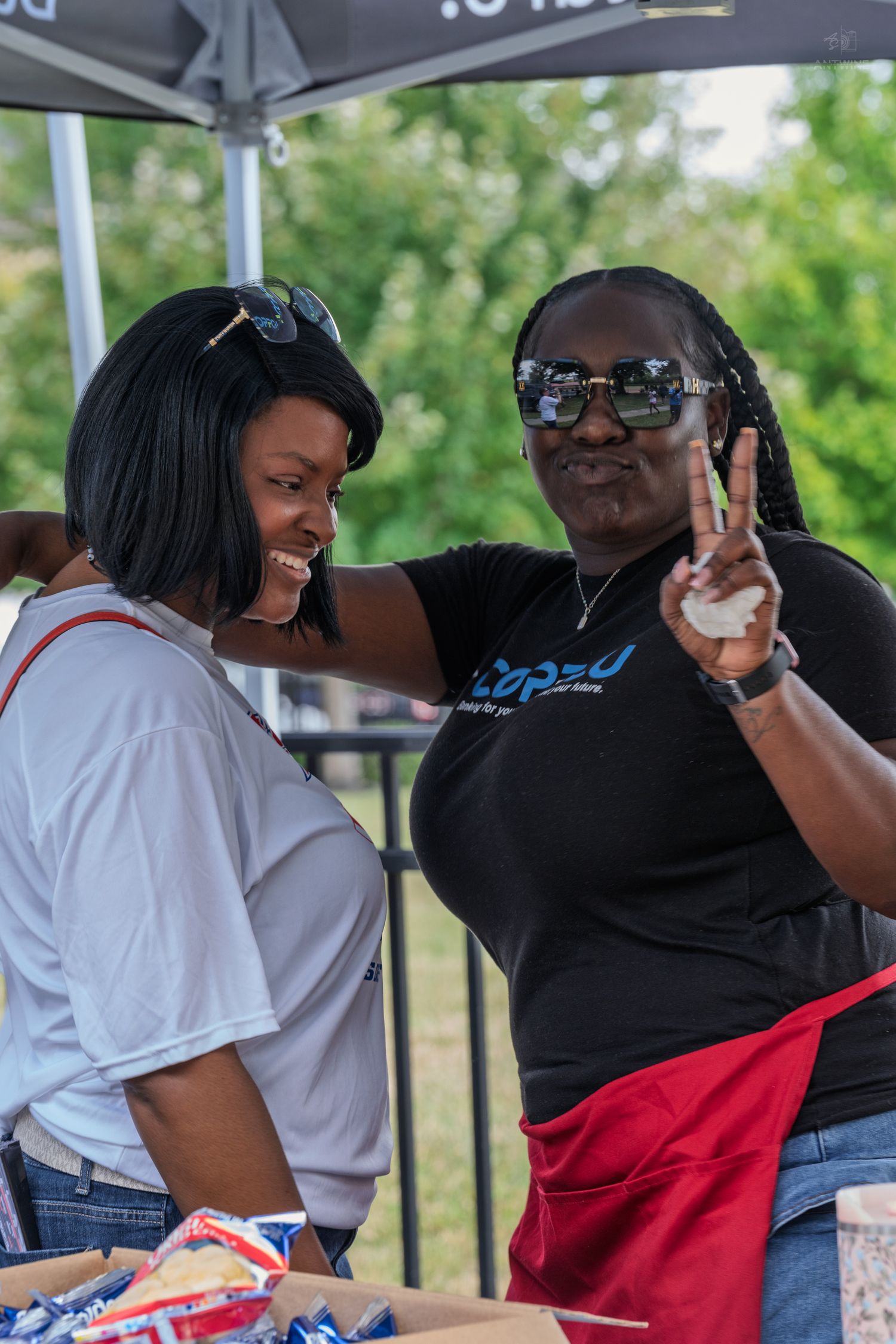 Two women smile and pose with a peace sign at an outdoor community food booth under a canopy.