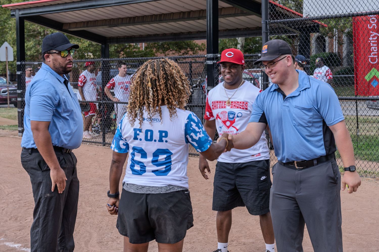 Players in FOP jerseys shaking hands with umpires near a dugout at an outdoor softball game.